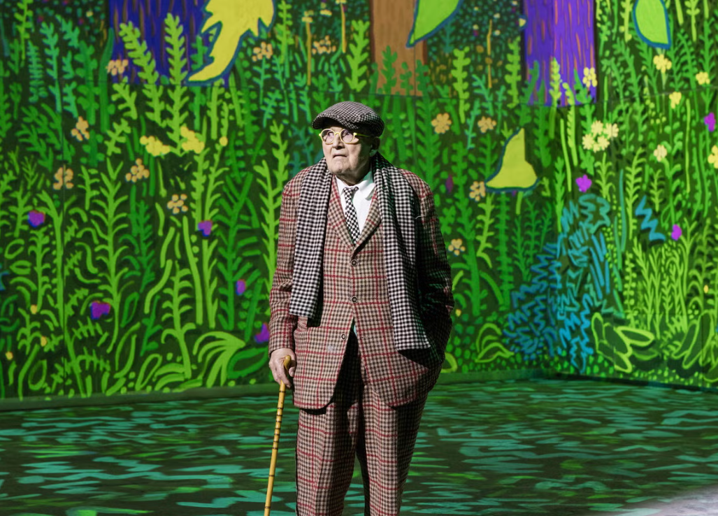 An elderly man dressed in a checkered suit and scarf, wearing a hat and glasses, standing on a stage with a colorful, cartoonish jungle background.