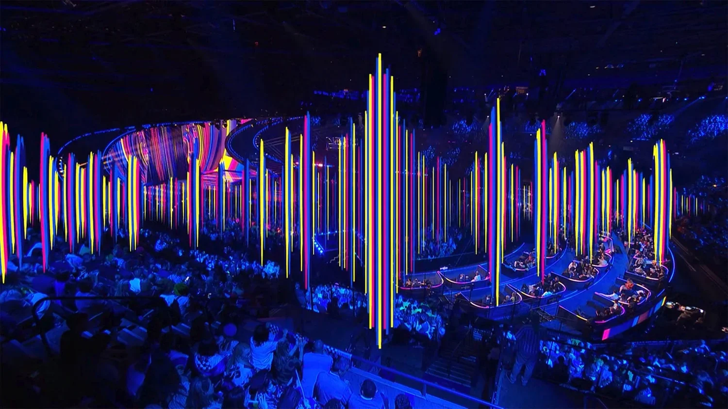 Dark indoor venue with colorful vertical neon lights and a crowd watching an event on a stage.