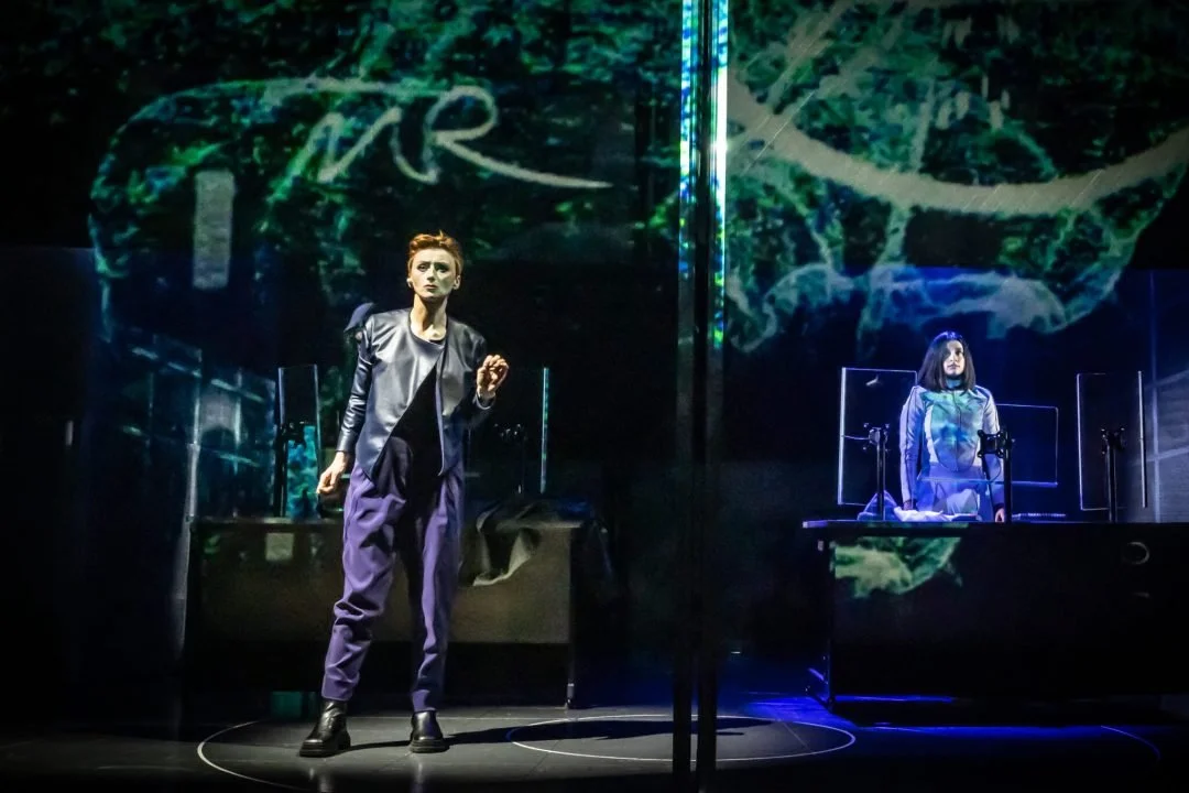 Stage scene with two women, one standing and one seated, with abstract digital art background.