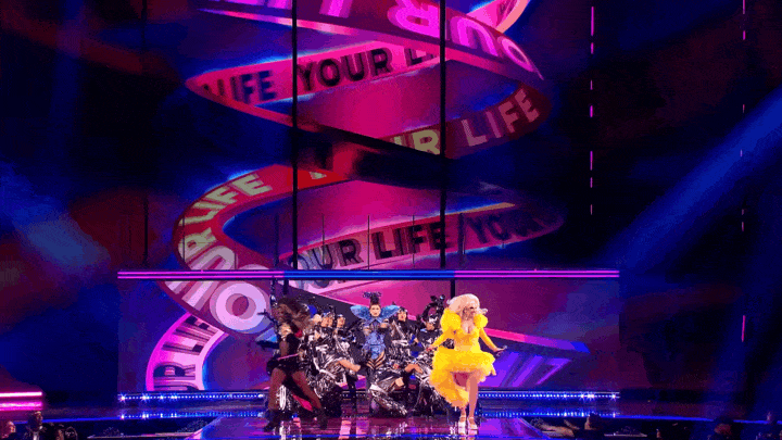 A stage with colorful lighting and a large screen in the background displaying the lyrics 'LIFE IN YOUR LIFE' in a spiral pattern. There is a woman with a large yellow dress singing in front of a group of dancers dressed in various costumes.