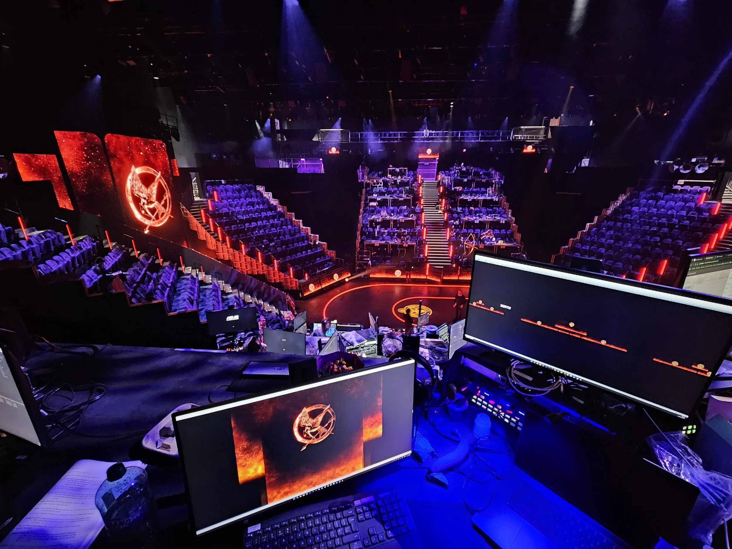 View of an empty theater or conference venue with purple seating, illuminated screen displays related to 'Hunger Games,' and a stage with stairs, as seen from a control booth with monitors and equipment.