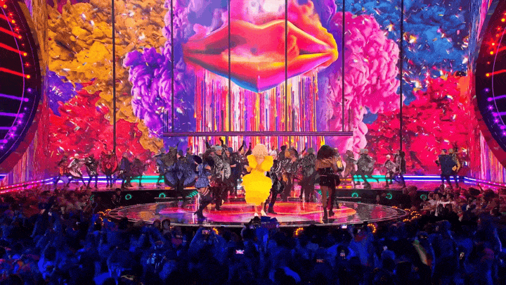 Live performance on stage featuring dancers and a performer in a yellow costume with a colorful, vibrant digital backdrop of flowers and an abstract face.