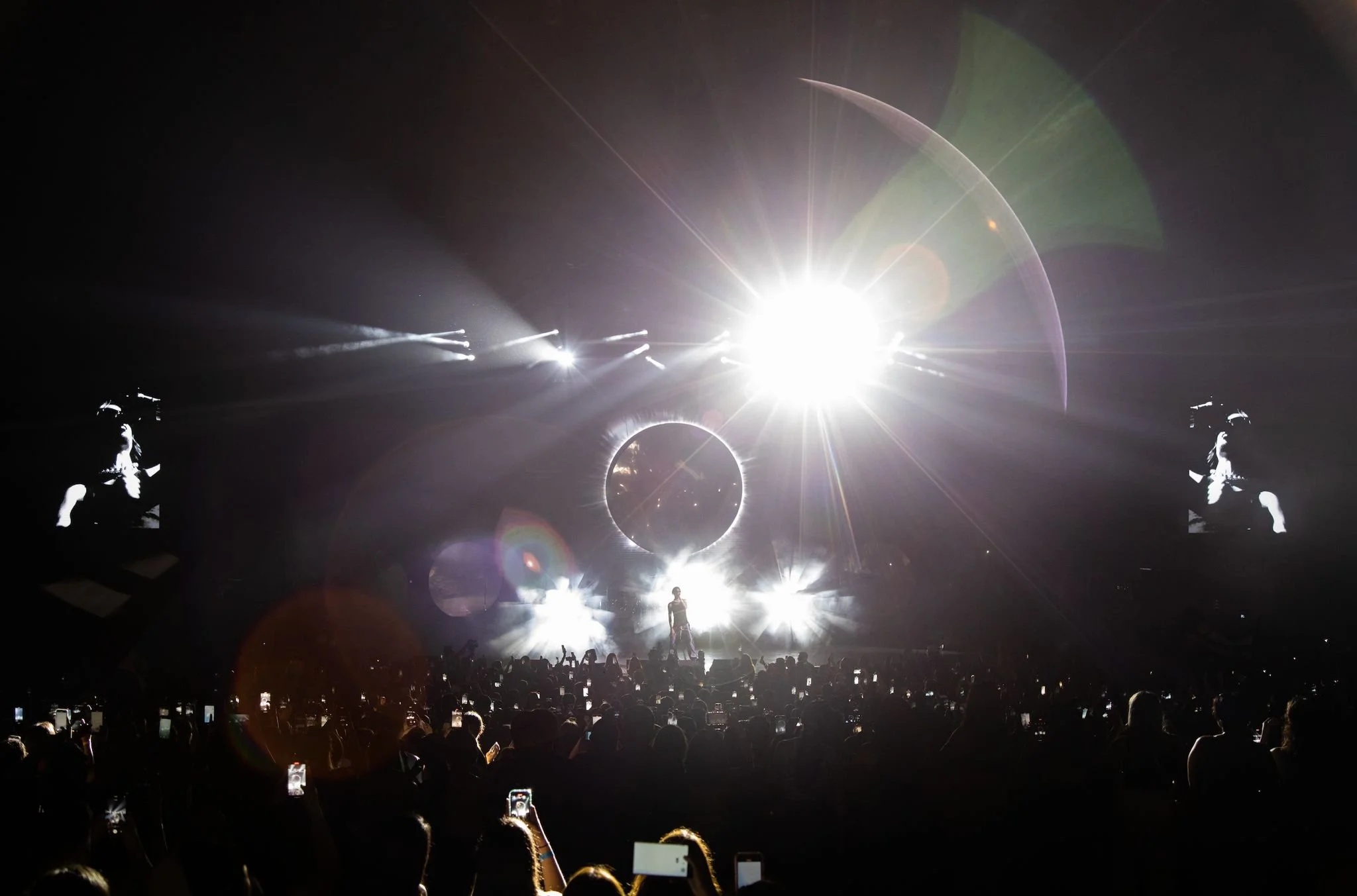 A concert scene with a performer on stage surrounded by bright lights, large circular screens, and an enthusiastic crowd capturing the moment with their phones.