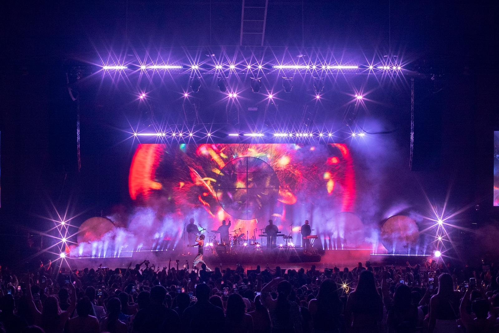 A live concert with a band performing on stage, vibrant purple and red lighting, and a large digital screen in the background showing abstract visuals. The audience is standing and watching the performance.