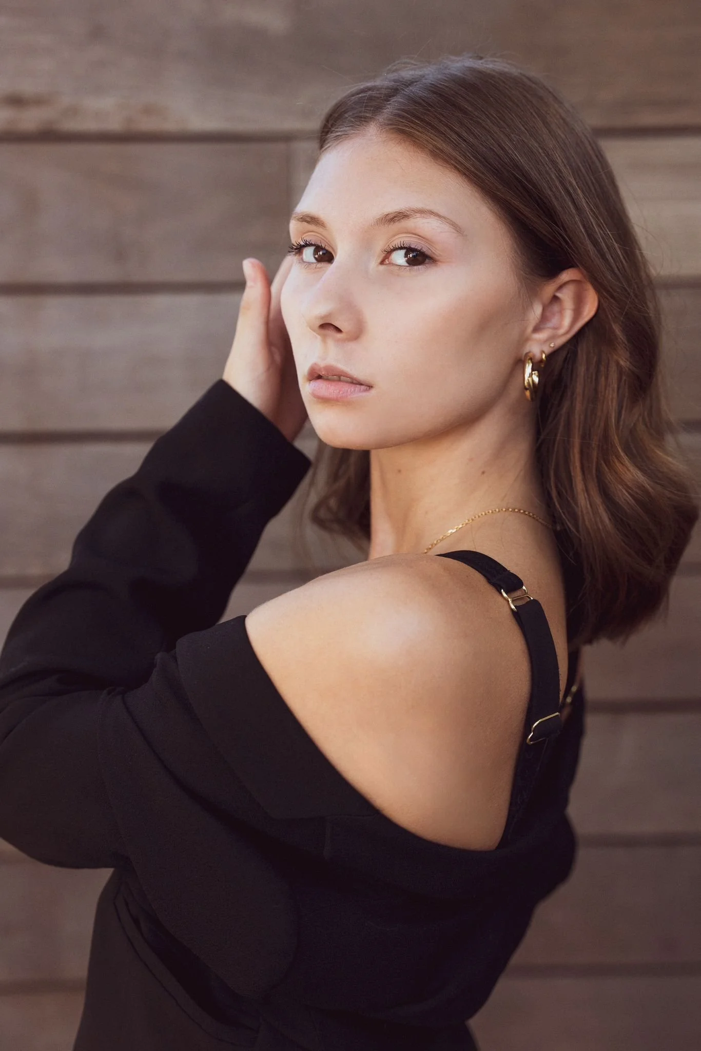 A woman with shoulder-length brown hair, wearing a black top with one shoulder exposed, gold earrings, and a gold necklace, standing against a wooden wall, looking at the camera with her hand touching her head.