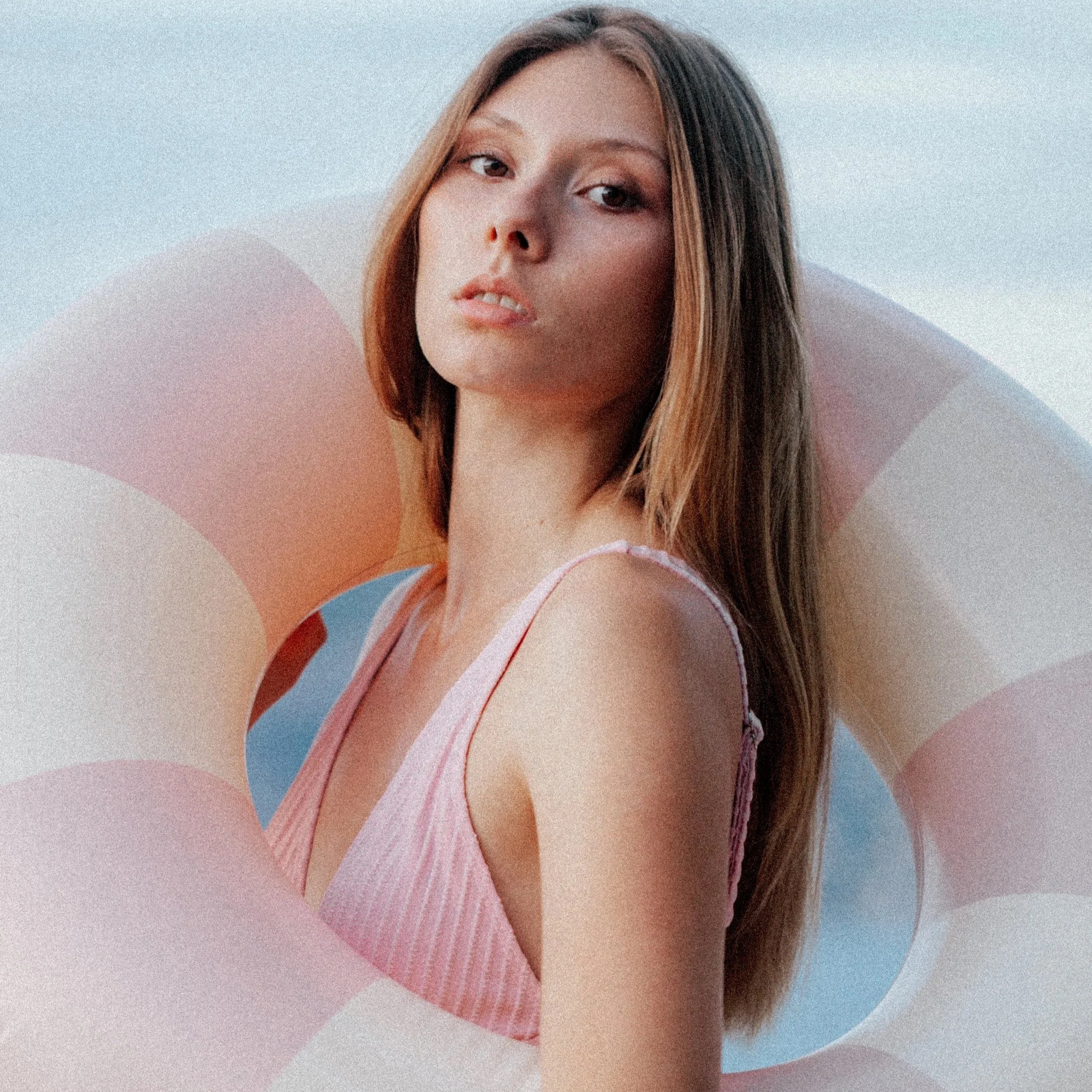 A young woman with long brown hair wearing a pink striped tank top looking at the camera, with a pastel-colored float in the background.