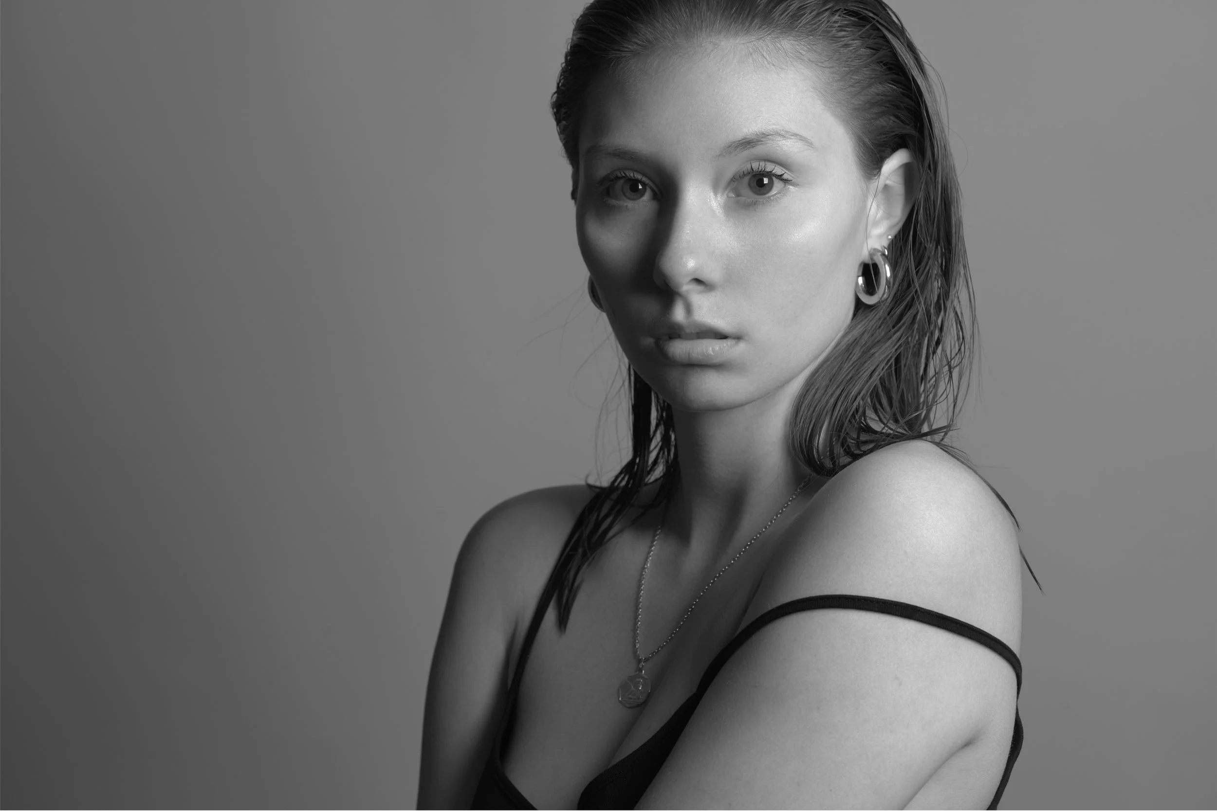 Black and white portrait of a young woman with wet hair, wearing earrings, a necklace, and a spaghetti strap top, looking into the camera.