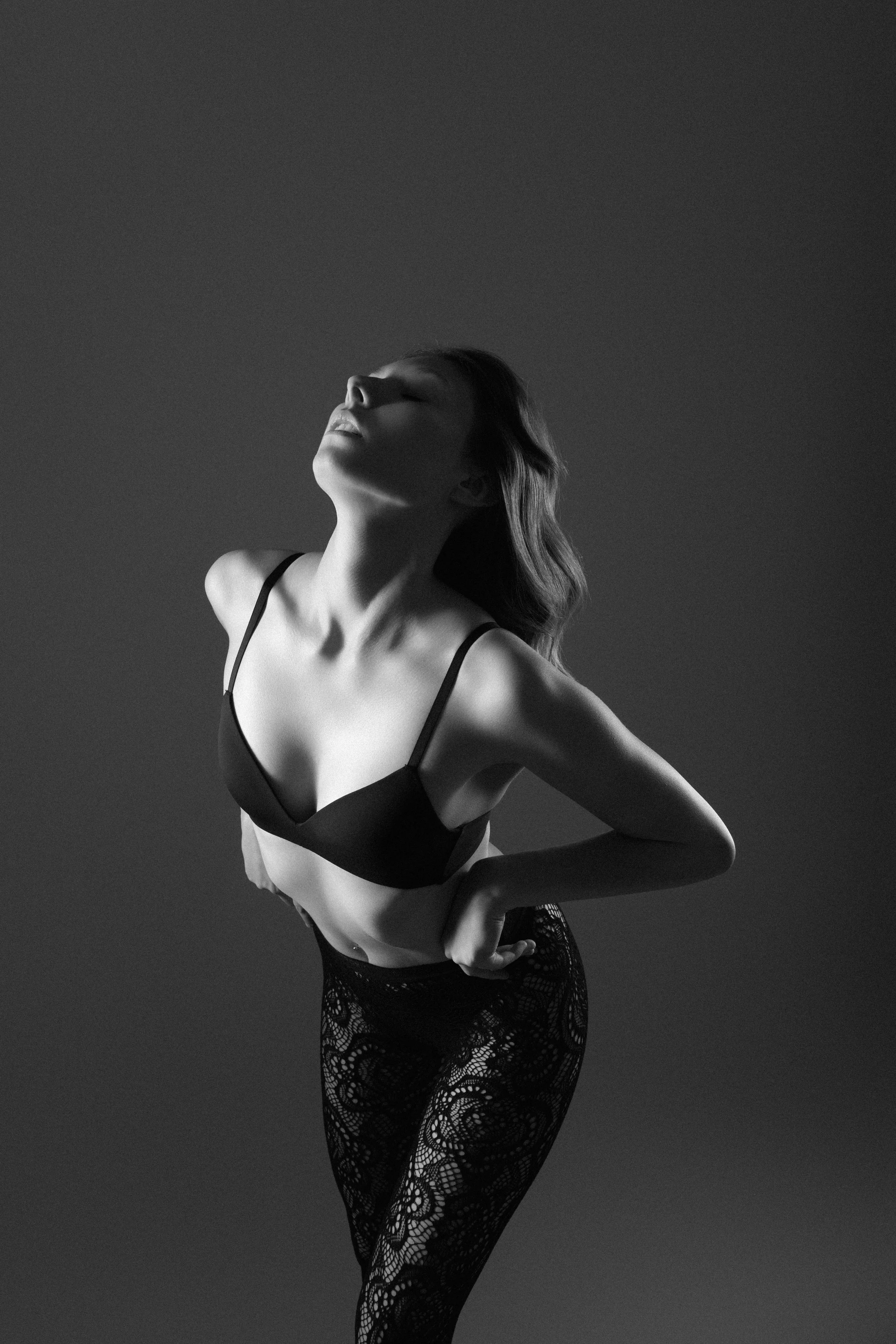 Black and white photo of a woman in a bra and lace leggings posing with her head tilted back and eyes closed.