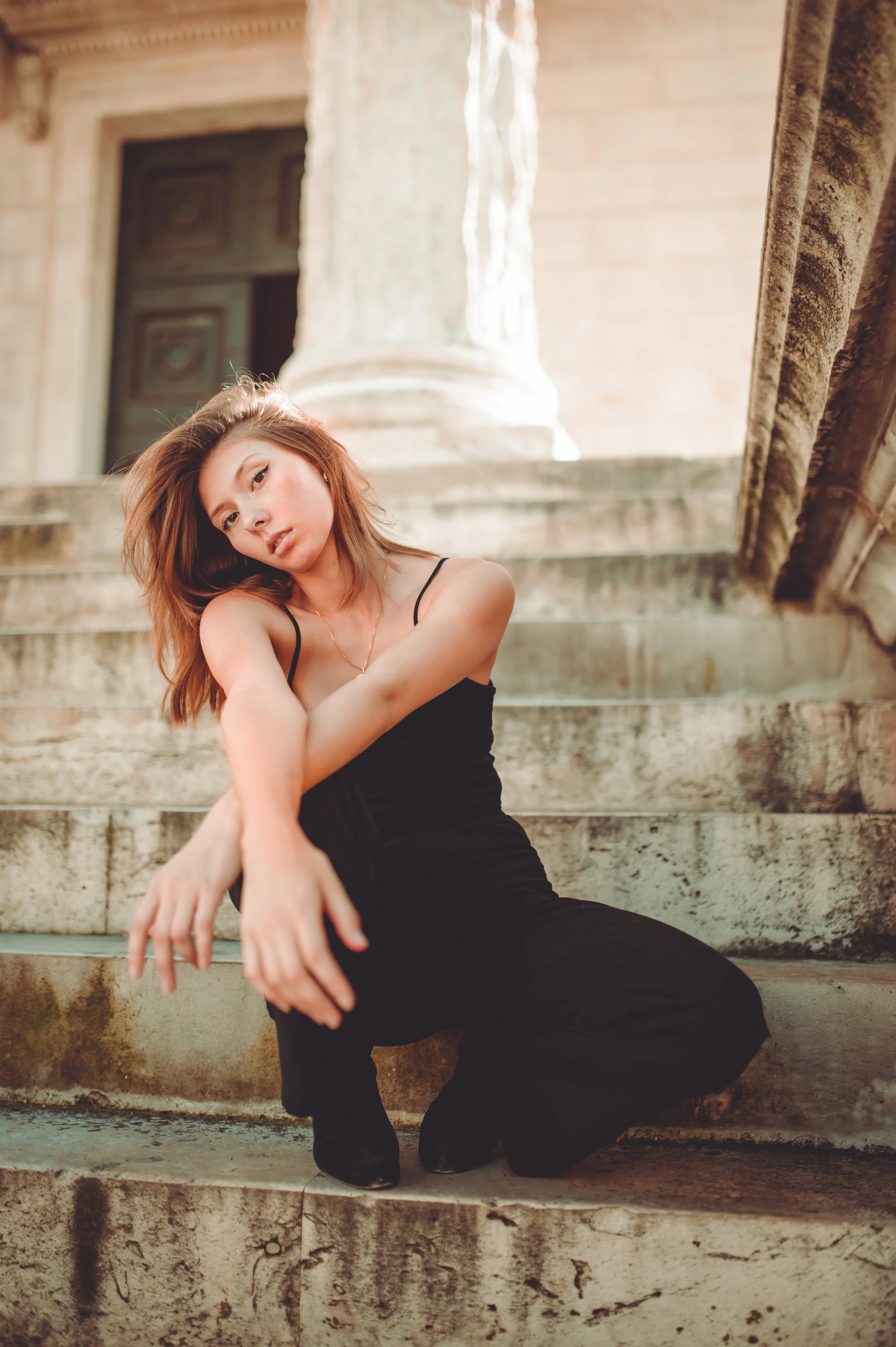A young woman with shoulder-length brown hair wearing a black dress sitting on weathered stone steps in front of a historic building with a large column and ornate door.