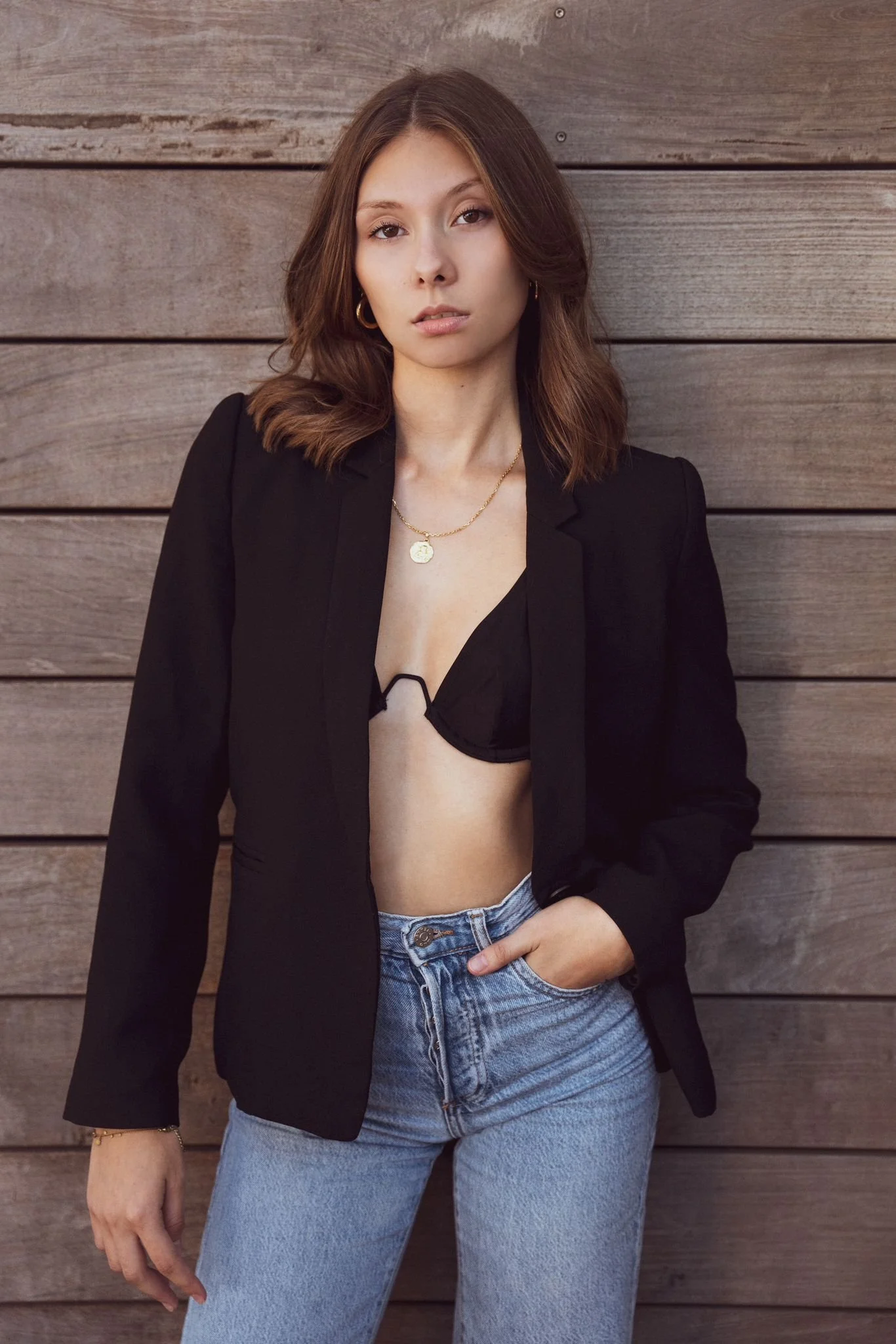 A young woman with medium-length brown hair, wearing a black blazer, a black bra, light blue jeans, gold hoop earrings, a gold necklace, and a bracelet, standing against a background of horizontal wooden planks.