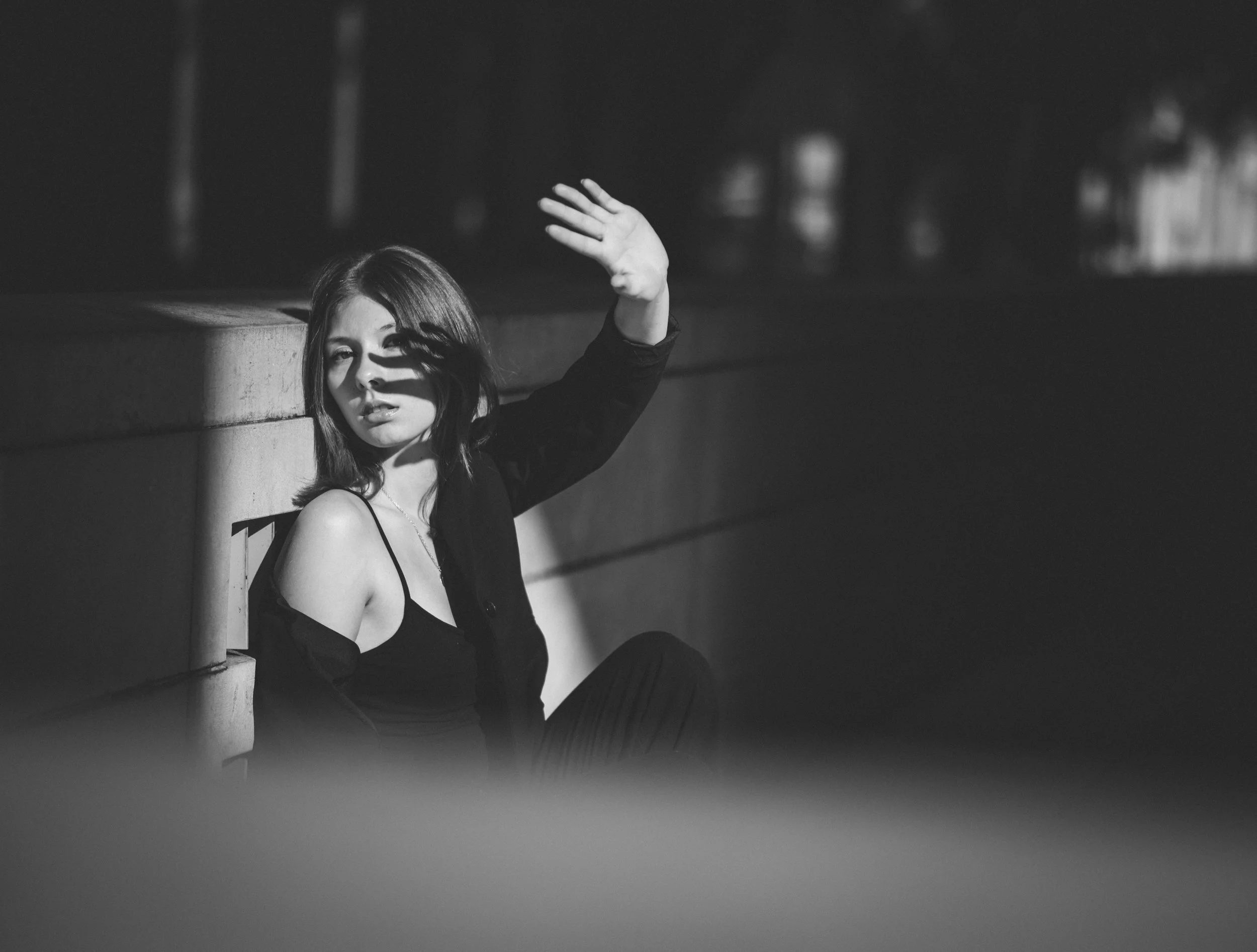 A black and white photo of a woman sitting outdoors on a ledge during nighttime. She has shoulder-length hair and is wearing a sleeveless top with a jacket. She is raising her right hand and appears to be shielding her eyes from the light as she look