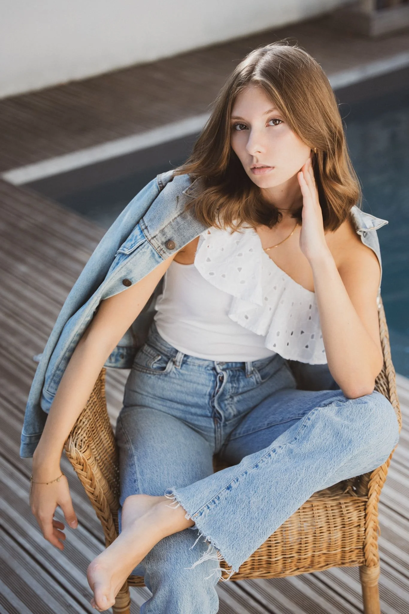A young woman with light brown hair sitting on a wicker chair on a wooden deck, wearing a white eyelet ruffle top, a denim jacket draped over her shoulders, and ripped jeans. She is barefoot and looking at the camera with one hand touching her neck.