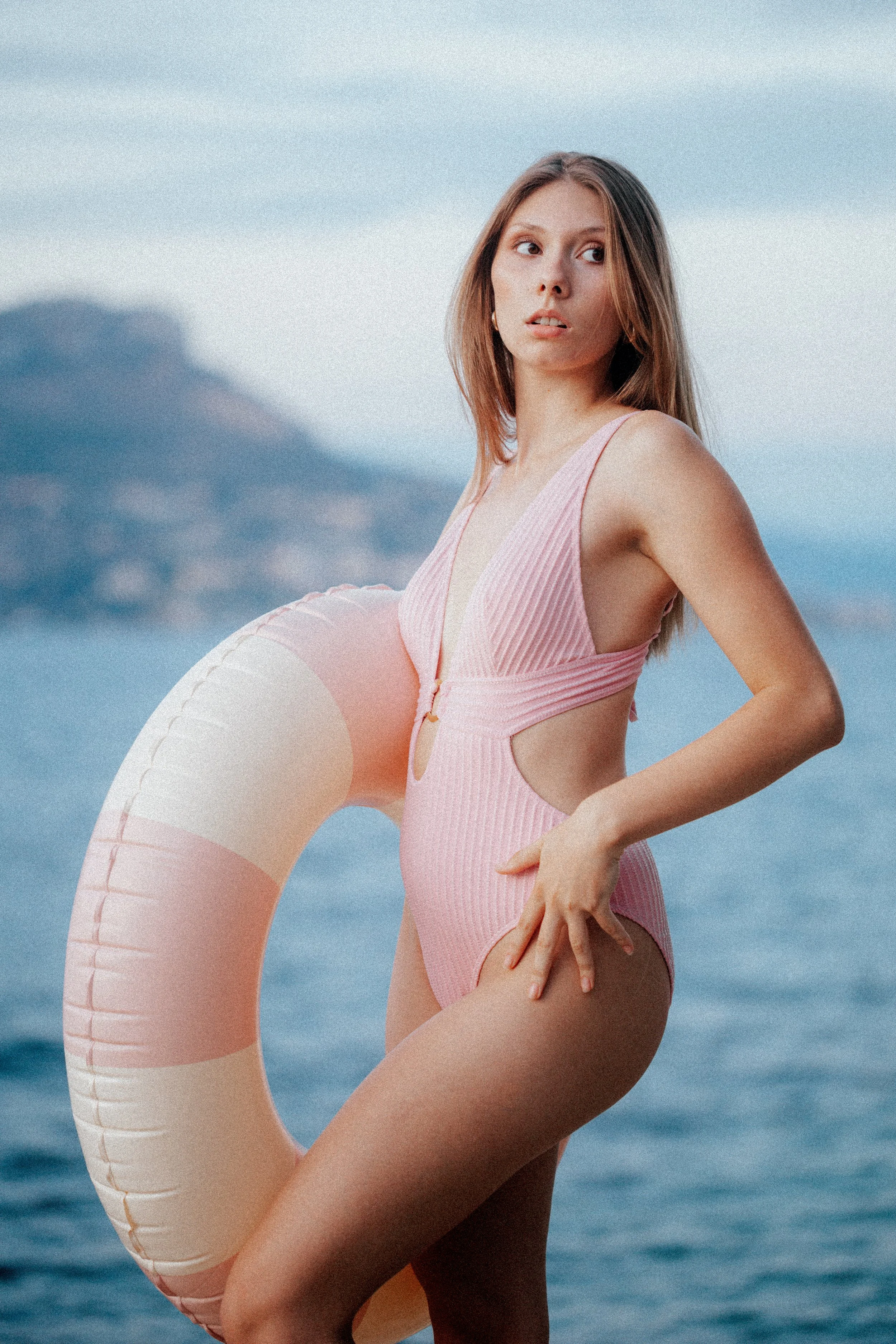 A woman wearing a pink swimsuit standing near the water, holding a large inflatable pool float shaped like a donut with a background of water and distant mountains.