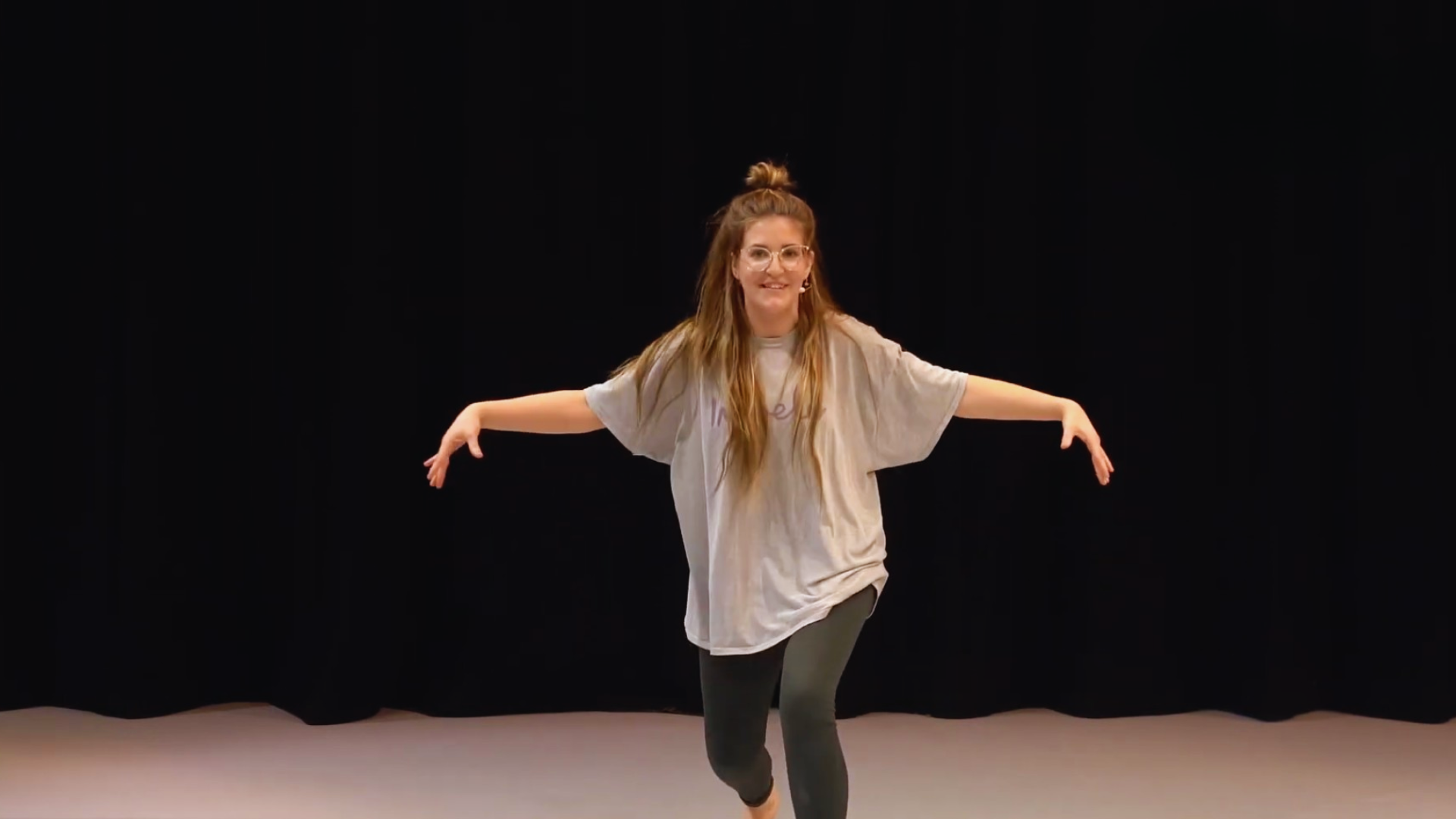 Young woman with long hair and glasses dancing on stage with her arms outstretched, wearing a loose fitting T-shirt and leggings.