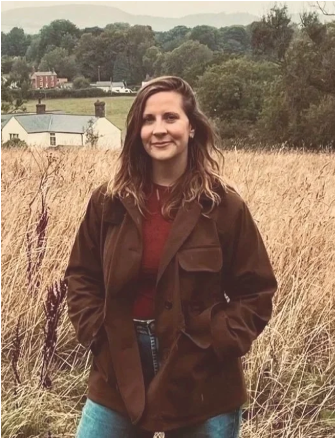 A woman standing in a field of tall grass with houses and trees in the background, smiling and wearing a brown jacket and red shirt.