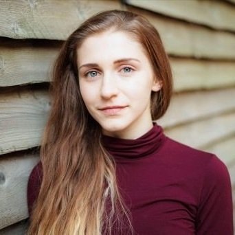  Image is of a female dancer from the shoulders up, it is a portrait photo. The background is grey wood and she is outside. Her top is red and her hair is light brown and long.  