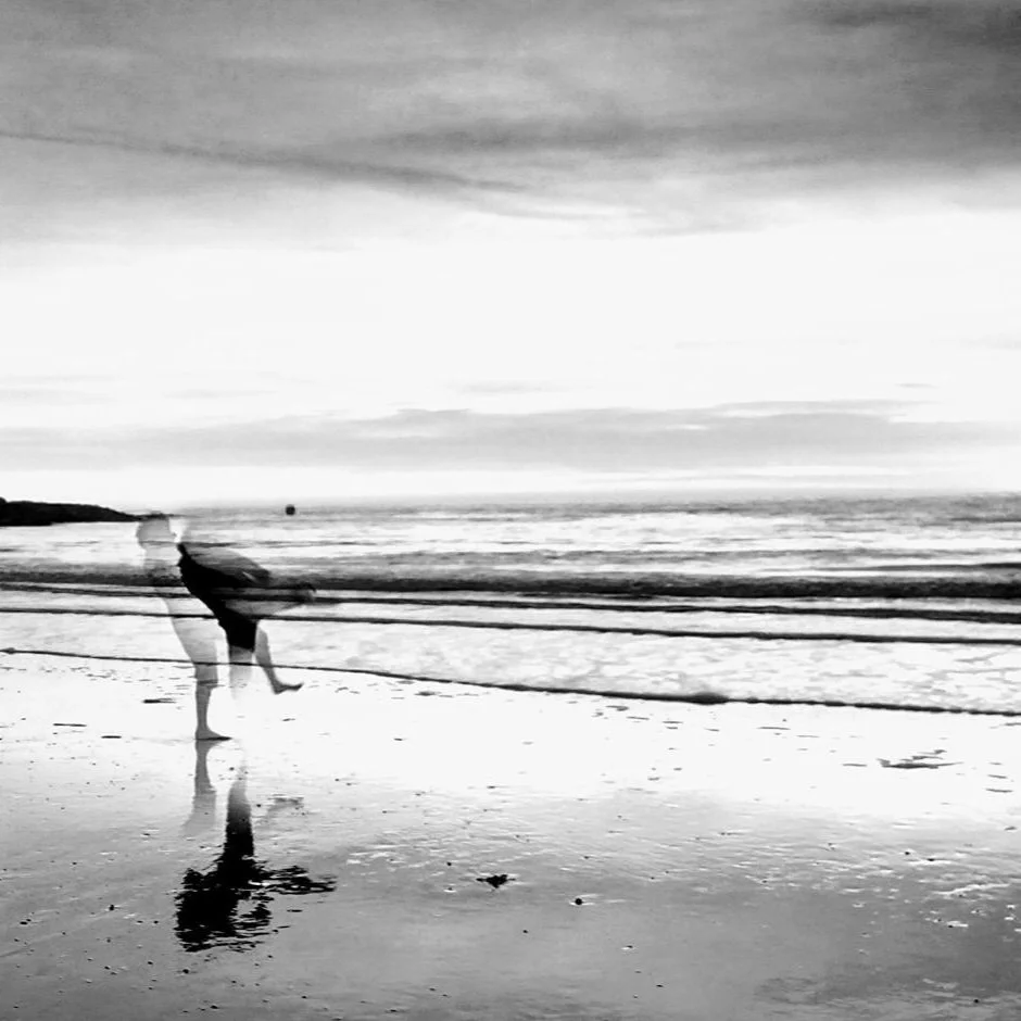  The image is black and white and mainly a landscape photograph of the beach with calm waves. The image shows the silhouette / faint outline of Fin and they are stood in the water with bare feet. 