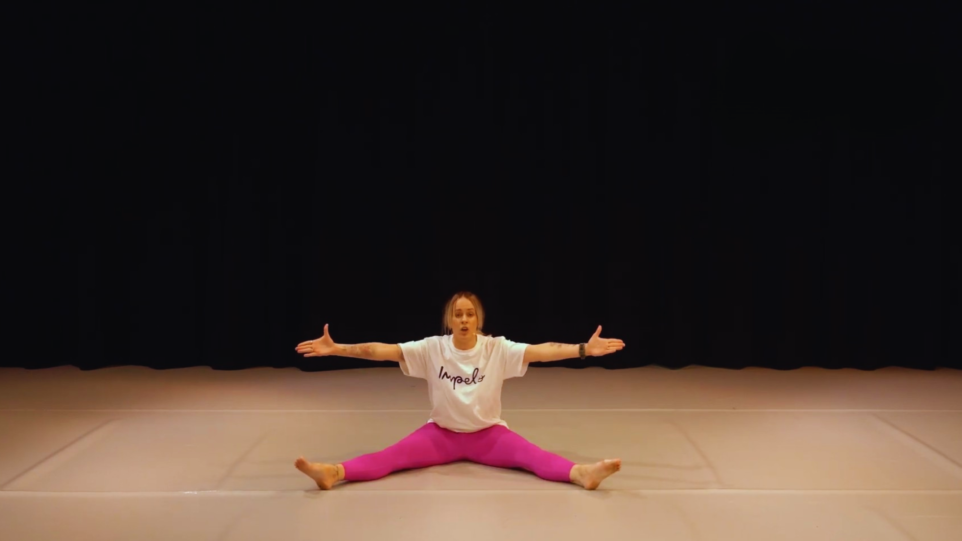 Woman sitting in a front split on a beige mat, with arms extended sideways, wearing a white t-shirt and pink leggings, against a black background.