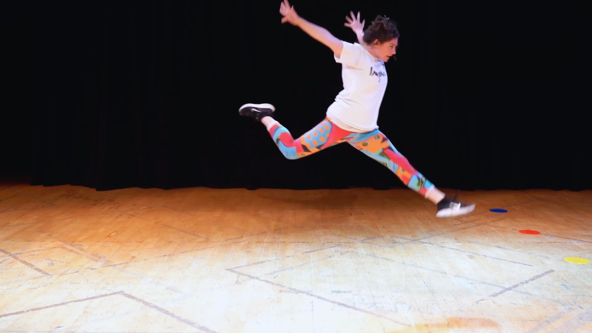 A woman in colorful leggings, a white t-shirt, and black sneakers is mid-air, jumping with arms outstretched and legs extended on a wooden floor with circular colored markers, against a black background.