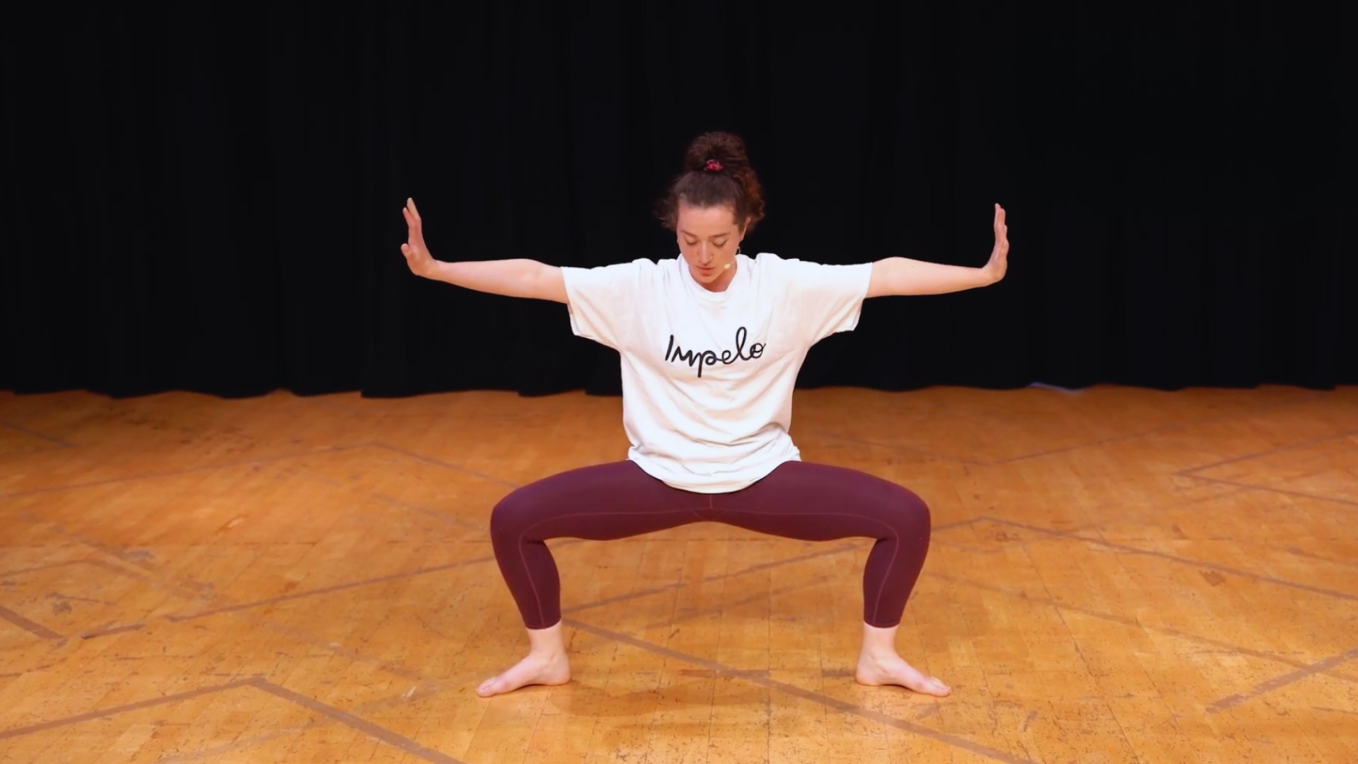 A woman performing a wide-legged squat with arms extended sideways inside a dance studio or stage with a wooden floor and black curtain background.