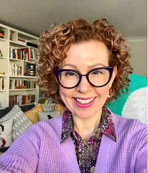 A woman with curly red hair and glasses taking a selfie in a room filled with bookshelves, wearing a purple cardigan and patterned shirt.