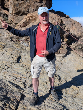 A man with a beard hiking on rocky terrain, wearing a red shirt, black jacket, beige shorts, and a white cap, with a blue sky background.