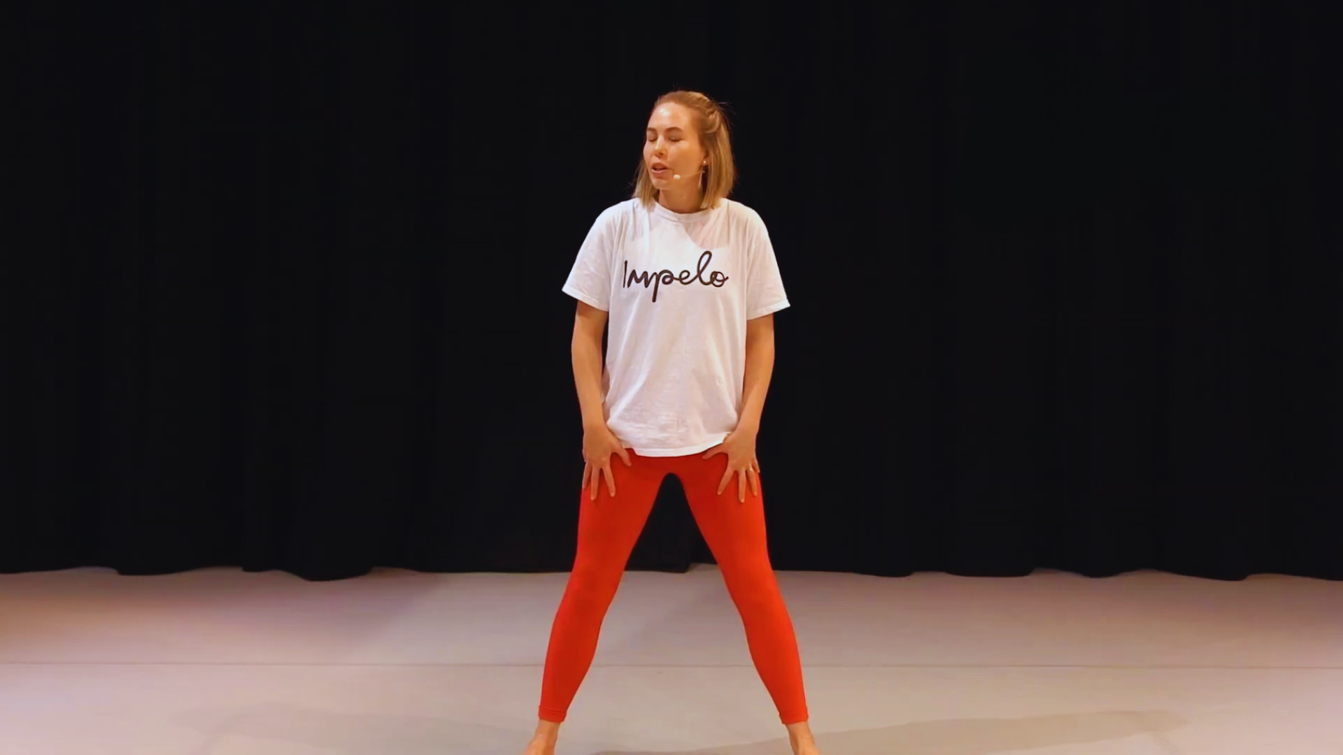 A woman with blonde hair in a beige top and red leggings standing with her legs apart and hands on her thighs on a stage with a black curtain background.