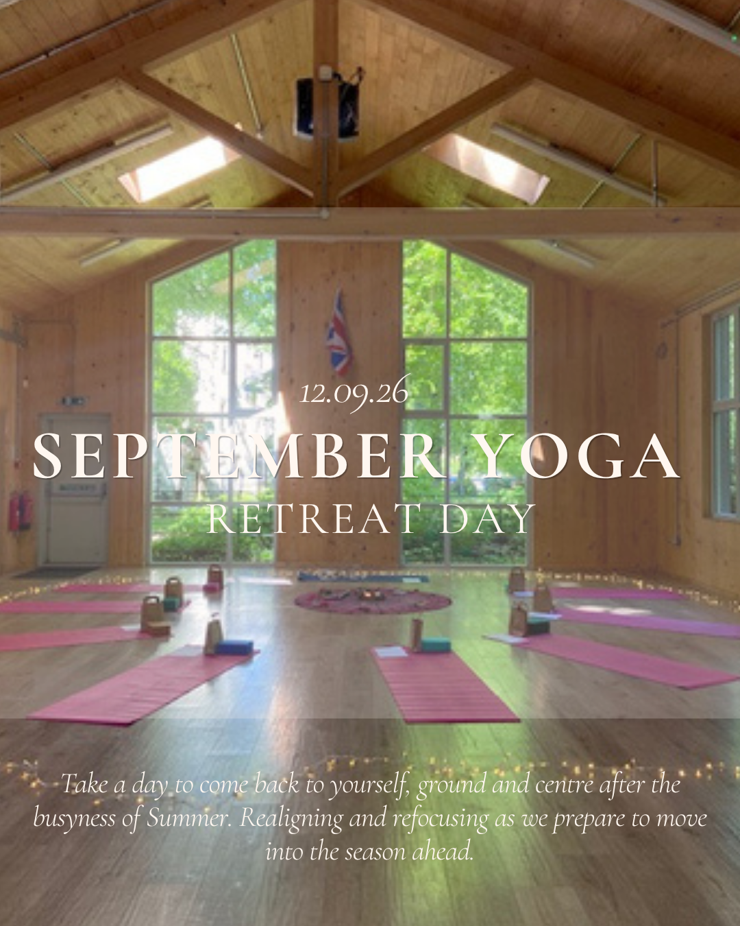 A spacious wooden yoga studio with floor mats, water bottles, small bags, and mats for meditation or yoga. Large windows provide natural light, and string lights are along the wall. The room appears set up for a yoga class or meditation session.