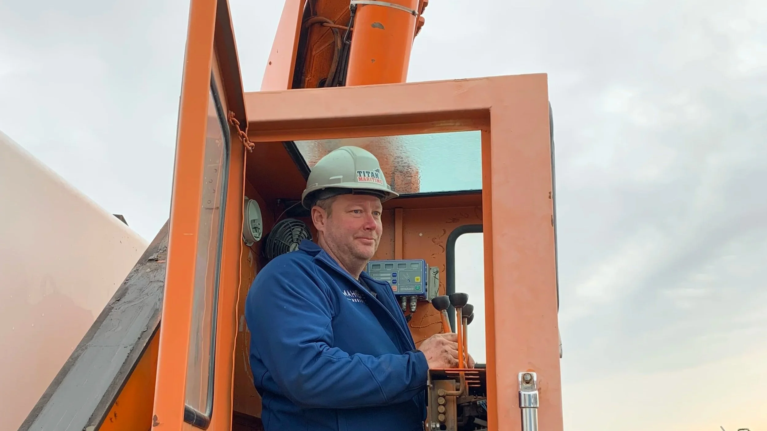 A construction worker in a blue jacket and a white safety helmet standing inside an orange vehicle's cab, operating controls against a cloudy sky background.