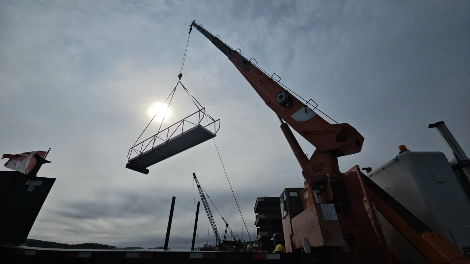 Construction crane lifting a large metal platform against a cloudy sky with the sun shining through.