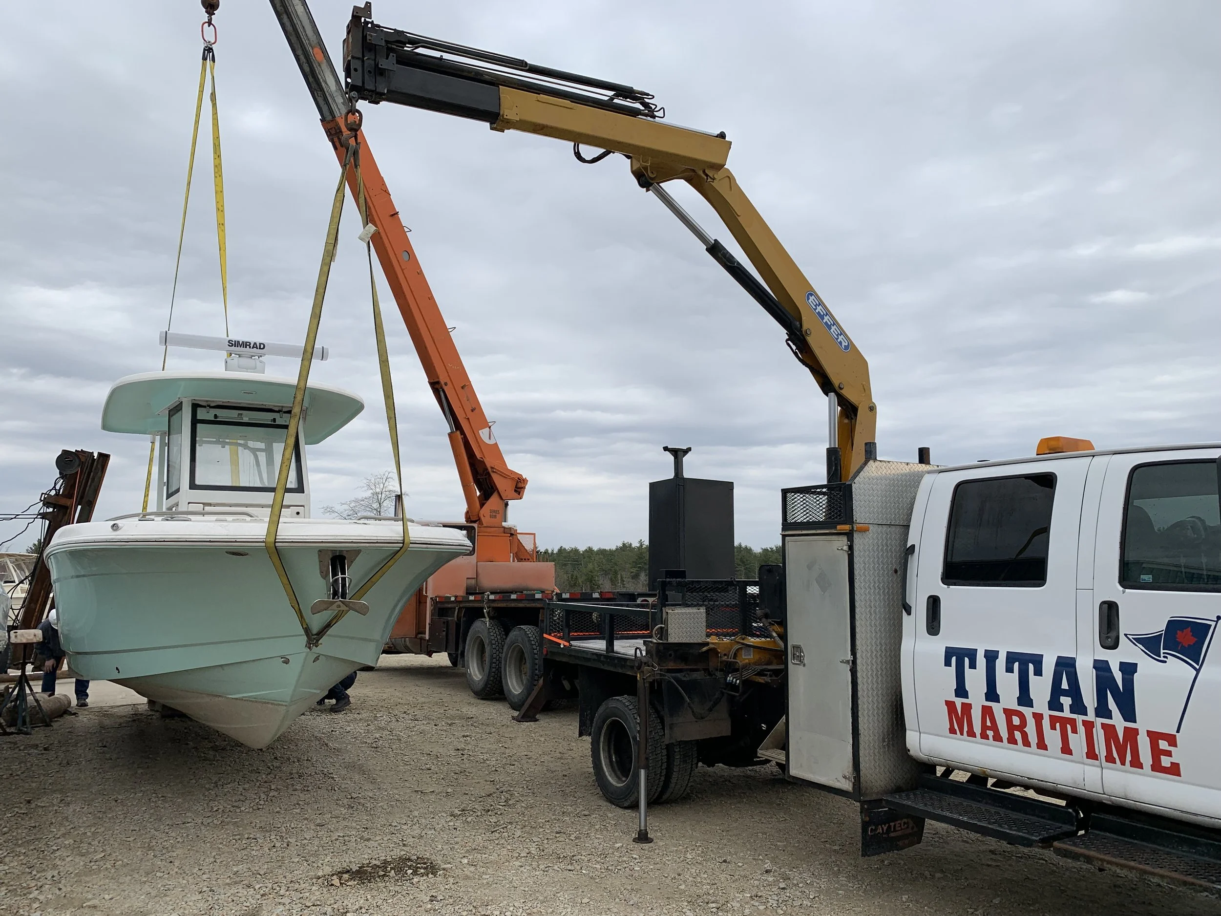 A boat is being lifted by a crane onto a truck at a marine service area, with the truck labeled "Titan Maritime." The scene is outdoors with a cloudy sky.