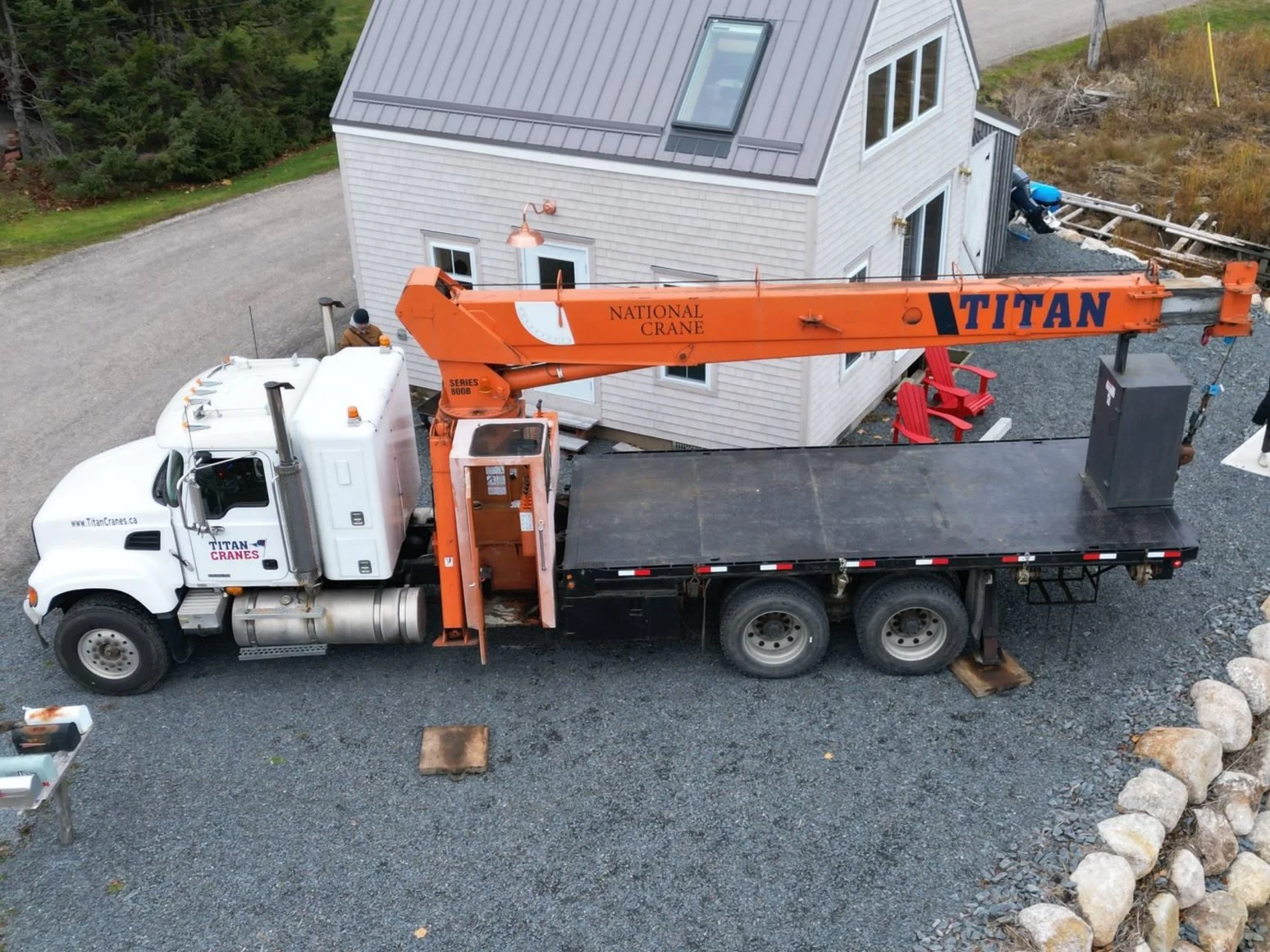 A white flatbed truck with an orange crane labeled 'Titan' parked in a gravel driveway next to a white house with a gray metal roof and large windows, with outdoor seating and some construction materials nearby.