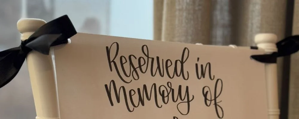 Personalised Reserved Sign, "Reserved in memory of" visible on the sign, attached to a white chair with black ribbon.