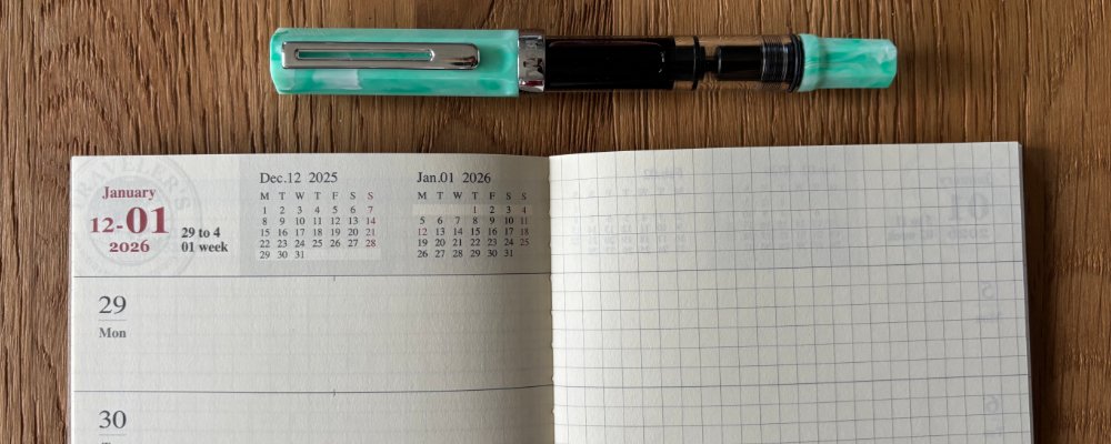 A fresh new diary insert lays open flat on a wooden work surface. A pale green fountain pen lays above it. Nothing is yet written on the pages.