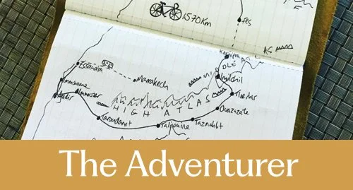 A notebook page with a hand-drawn map or route, including sketches of mountains and labels of locations, with a mustard banner at the bottom reading 'The Adventurer'.