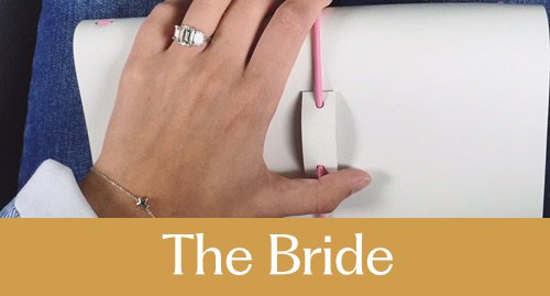 Close-up of a hand with an engagement ring, resting on a white leather journal, with a pink elastic, and a mustard band at the bottom with the text "The Bride."