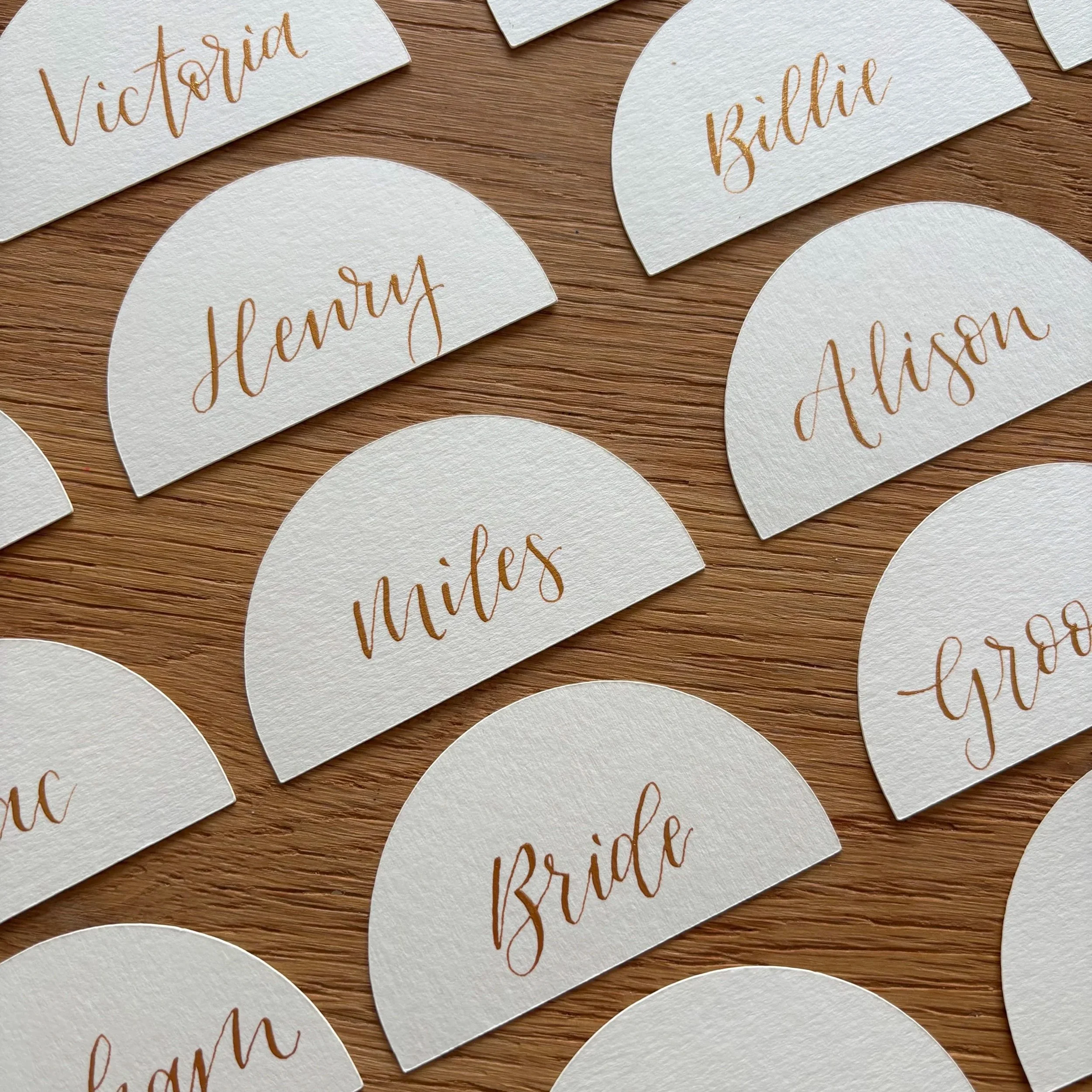 Off white semi circle cut place name cards, names written in gold modern calligraphy. Names - Victoria, Henry, Billie, Miles, Alison, Bride, Groom