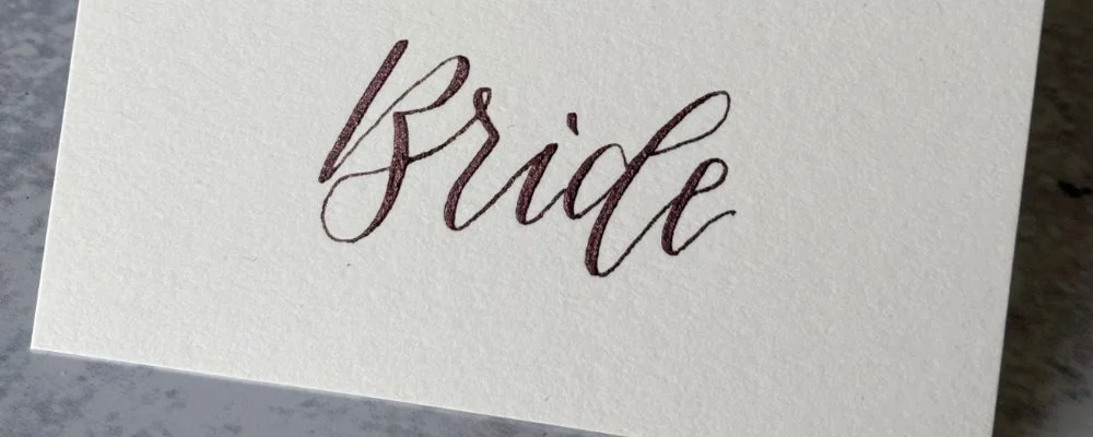 A folded place card, off white tented style, with the word "Bride" beautifully hand lettered in metallic chocolate brown modern calligraphy