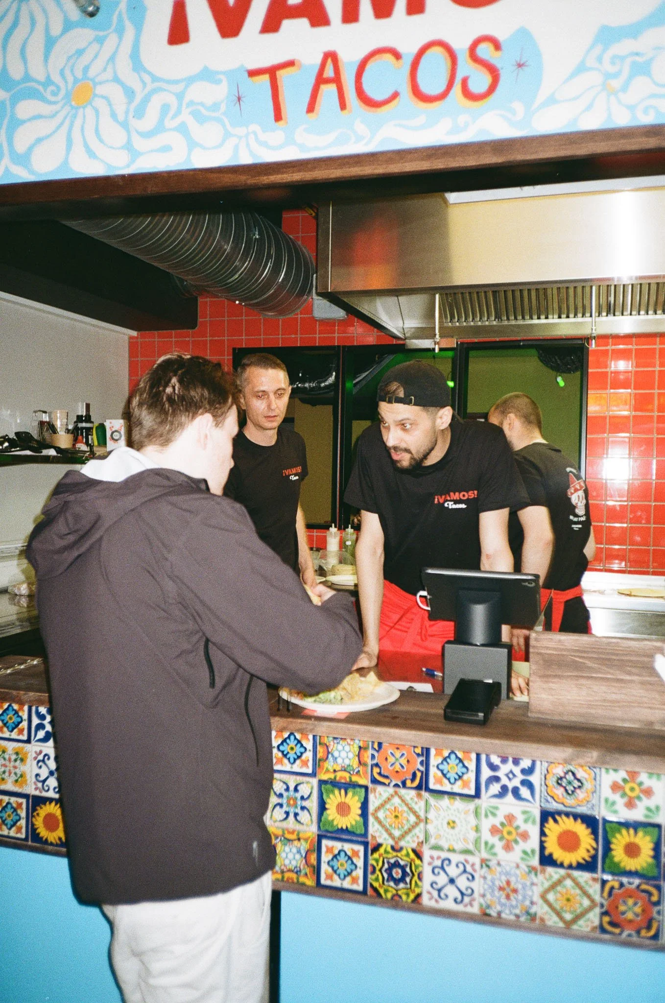 En kunde bestiller mat ved bardisken til en restaurant som serverer tacos. Personalet snakker med kunden, mens de står foran en fargerik vegg med mønstrede fliser.