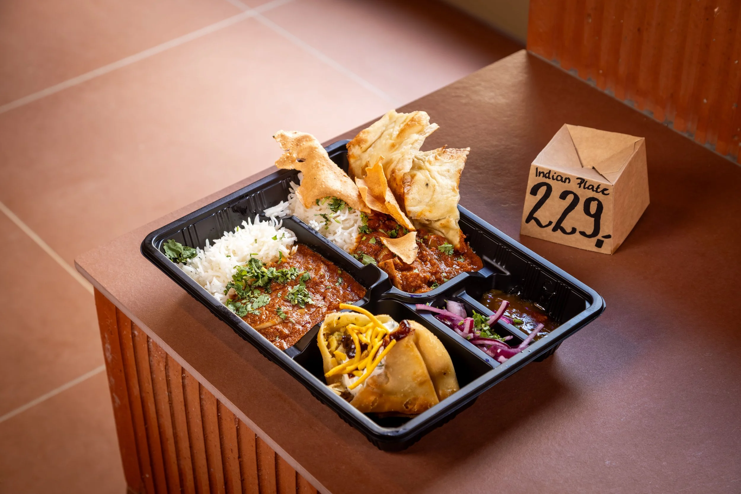 En plastboks med indisk mat på en bordplate, inkludert ris, curry, naanbrød, og ulike tilbehør.