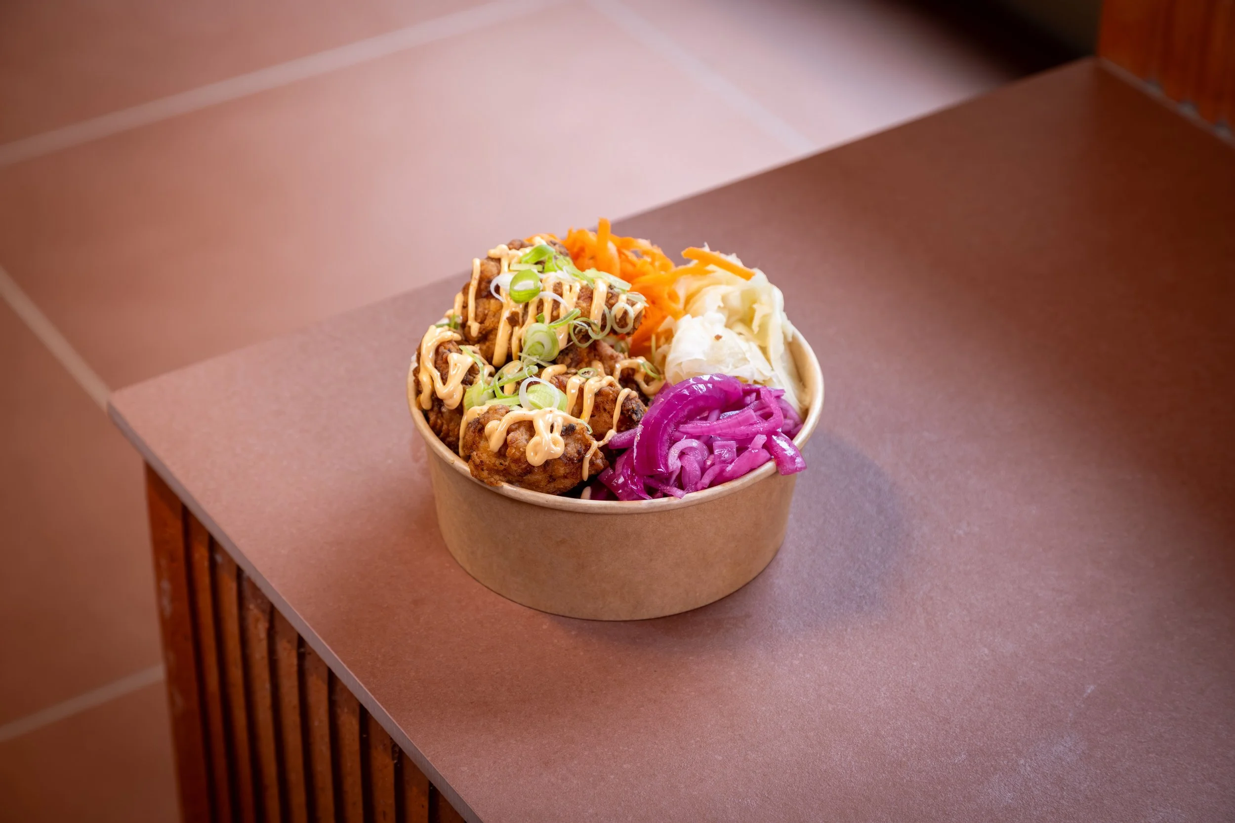 En pappkopper med ulike typer fylt og pyntet taco, inkludert kjøttboller, guacamole, gulrot, ost, og rødkål, plassert på et bord med trepanel og en lys rosa bordflate.