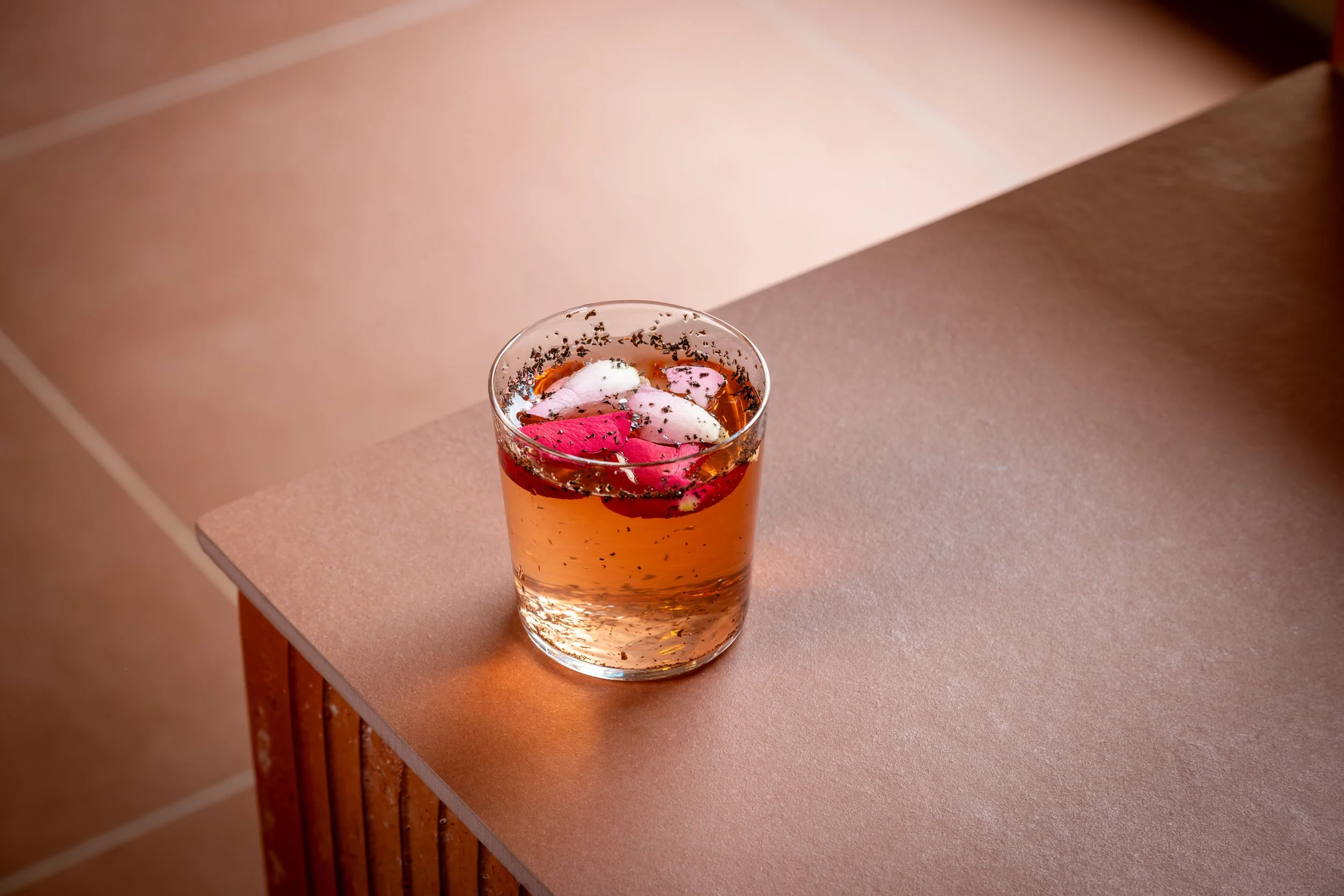 Et glass med en lyserosa drikkes med rosa og hvite marsipan- eller sukkertopper og litt svart krydder eller kanel på toppen, plassert på en bordflate.