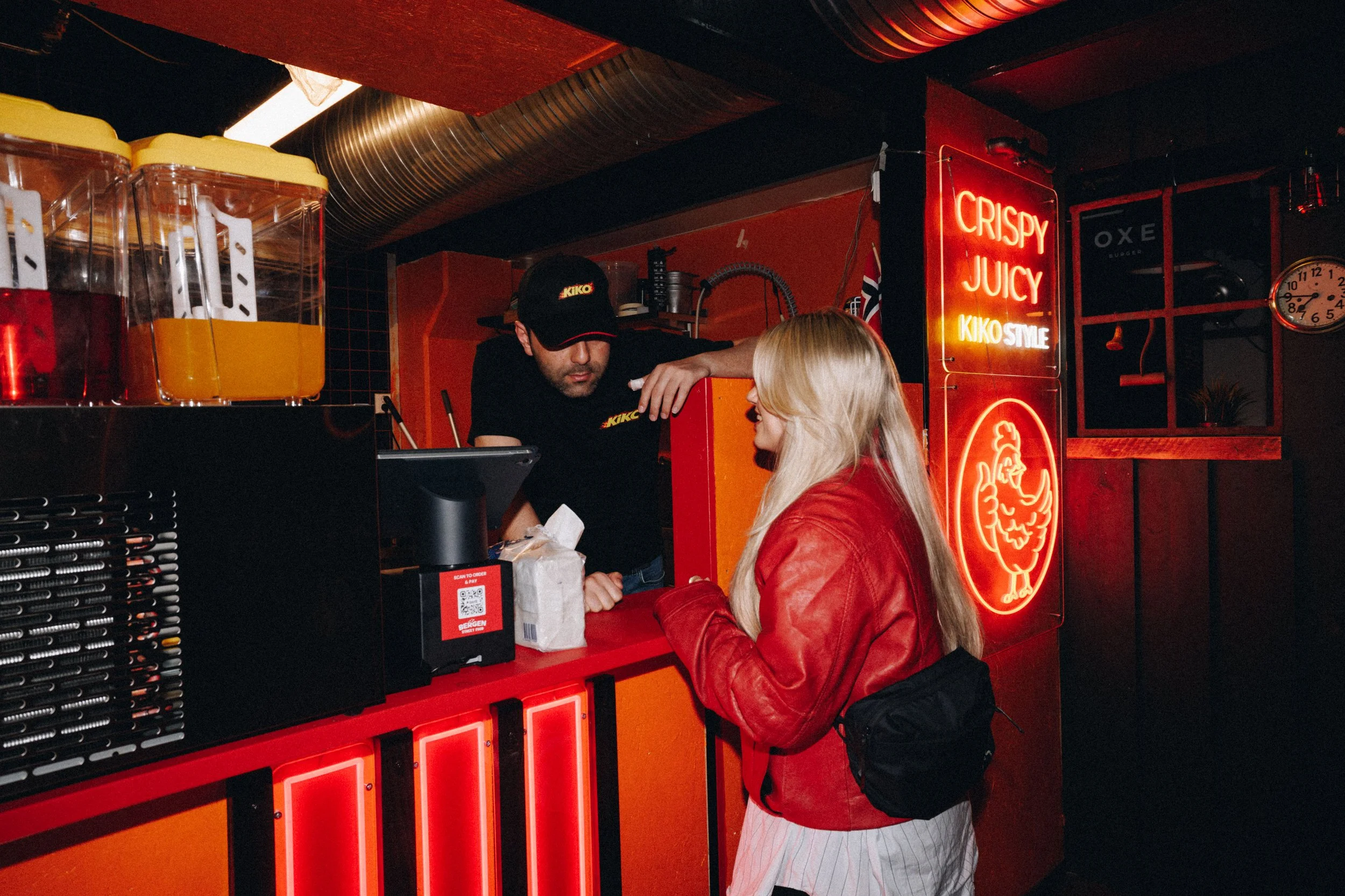 En kvinne i rød skinnjakke og hvit kjole kjøper mat fra en garçon bak disken på en restaurant eller burgerbar med røde og svarte interiørdetaljer og lysende skilt som sier "Crispy Juicy Kiko Style".