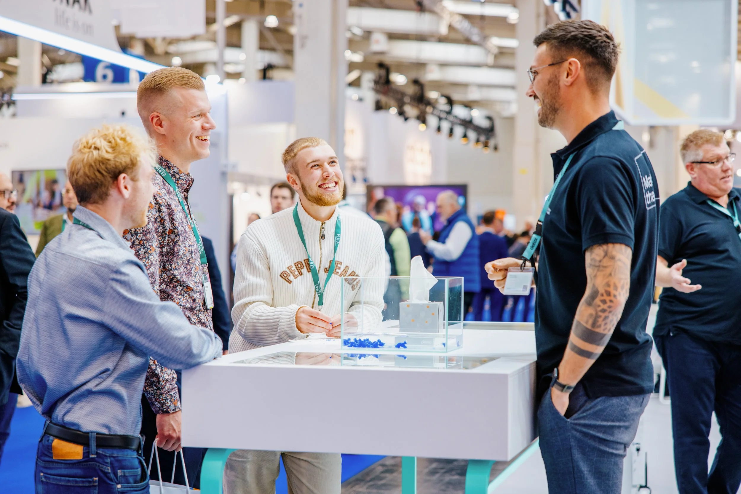 Vier junge Männer sprechen bei einer Messe oder Kongress. Sie lachen und unterhalten sich vor einem weißen Ausstellungstisch mit einer transparenten Box.
