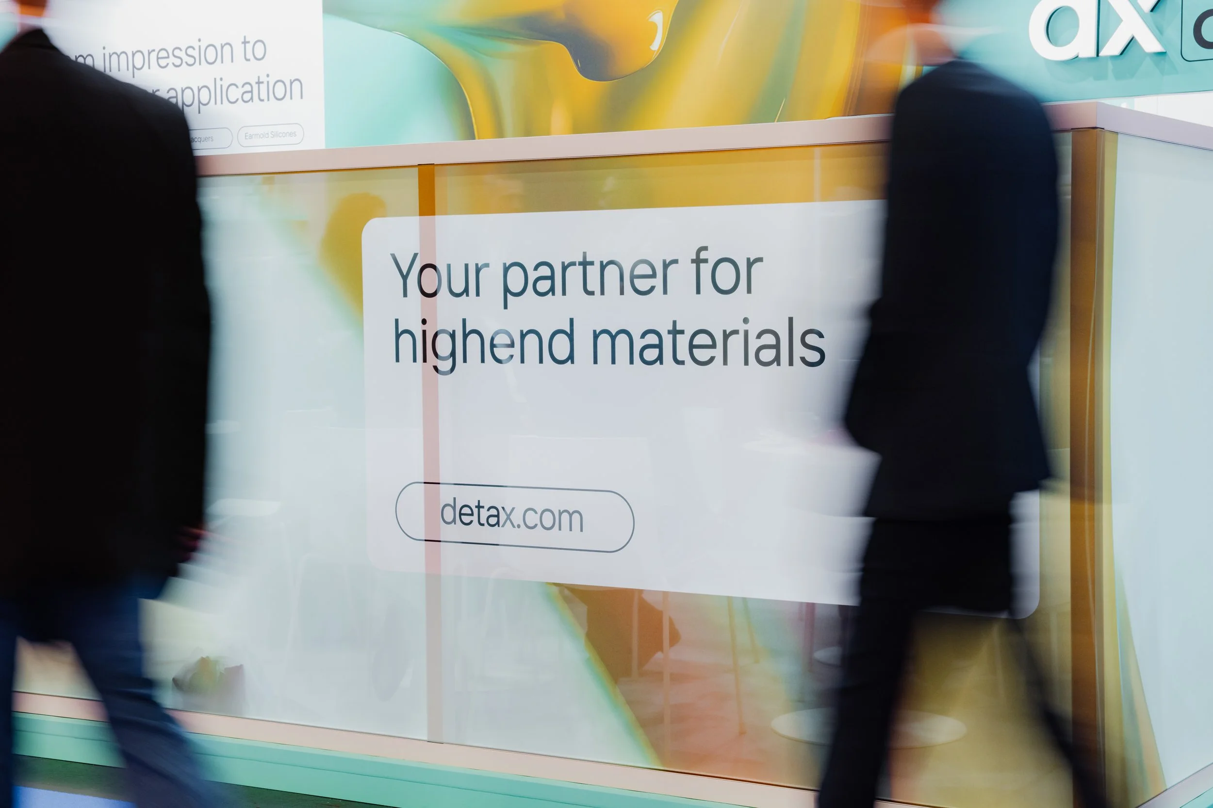 Werbung auf einer Messe mit großem weißen Schild, das sagt: 'Your partner for highend materials' und die Website 'detax.com'. Zwei Personen gehen vorbei, unscharf im Vordergrund.