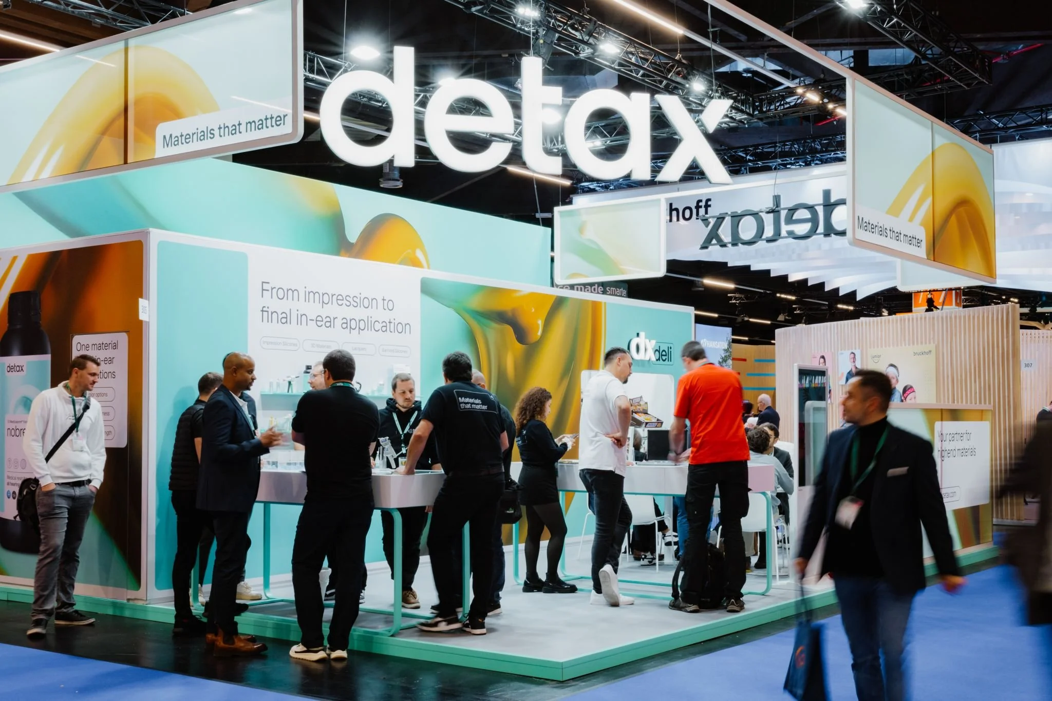 Ausstellungsstand bei einer Messe mit dem Logo 'detax' und mehreren Menschen, die sich informieren, einige im Gespräch, andere am Stand.