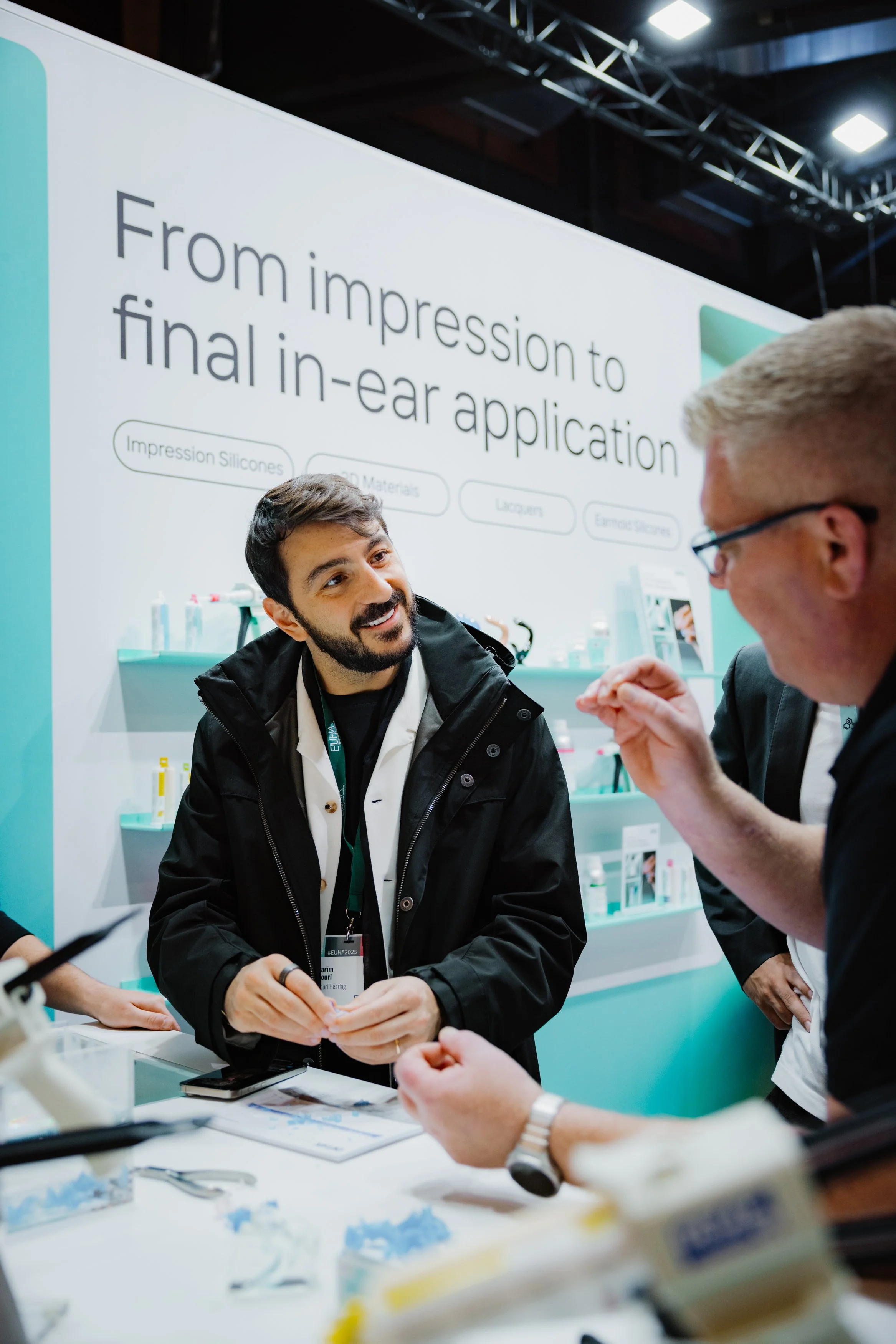 Zwei Männer unterhalten sich bei einem Fachkongress oder Messe, im Hintergrund ein großes Banner mit dem Text 'From impression to final in-ear application' und weiteren Stichpunkten.
