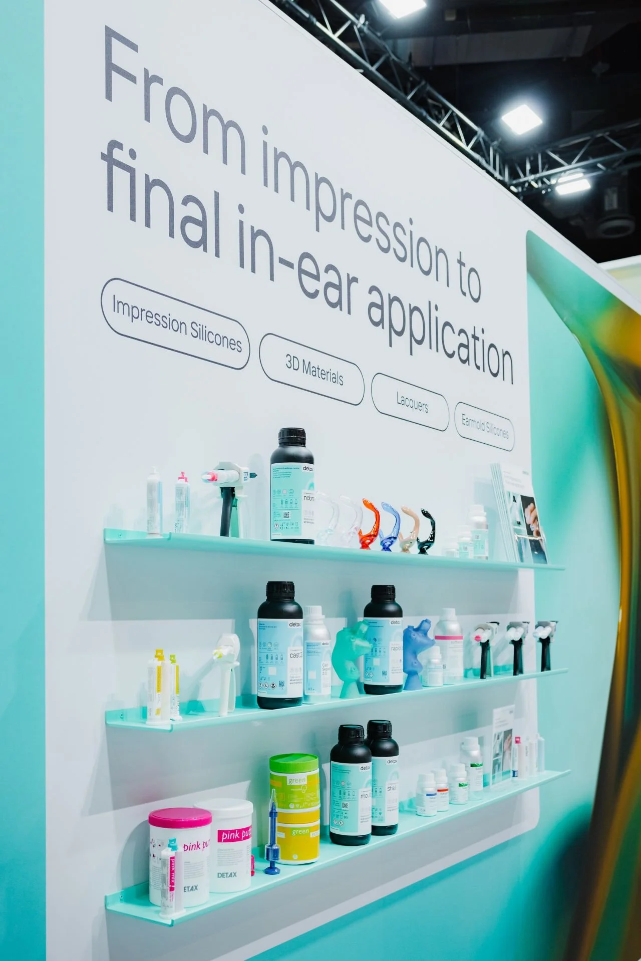 Präsentationswand mit Produkten für Final in-ear-Anwendungen, darunter Silikoneindrucke, 3D-Materialien, Lacke und Ohrstöpsel, auf mehreren Regalböden