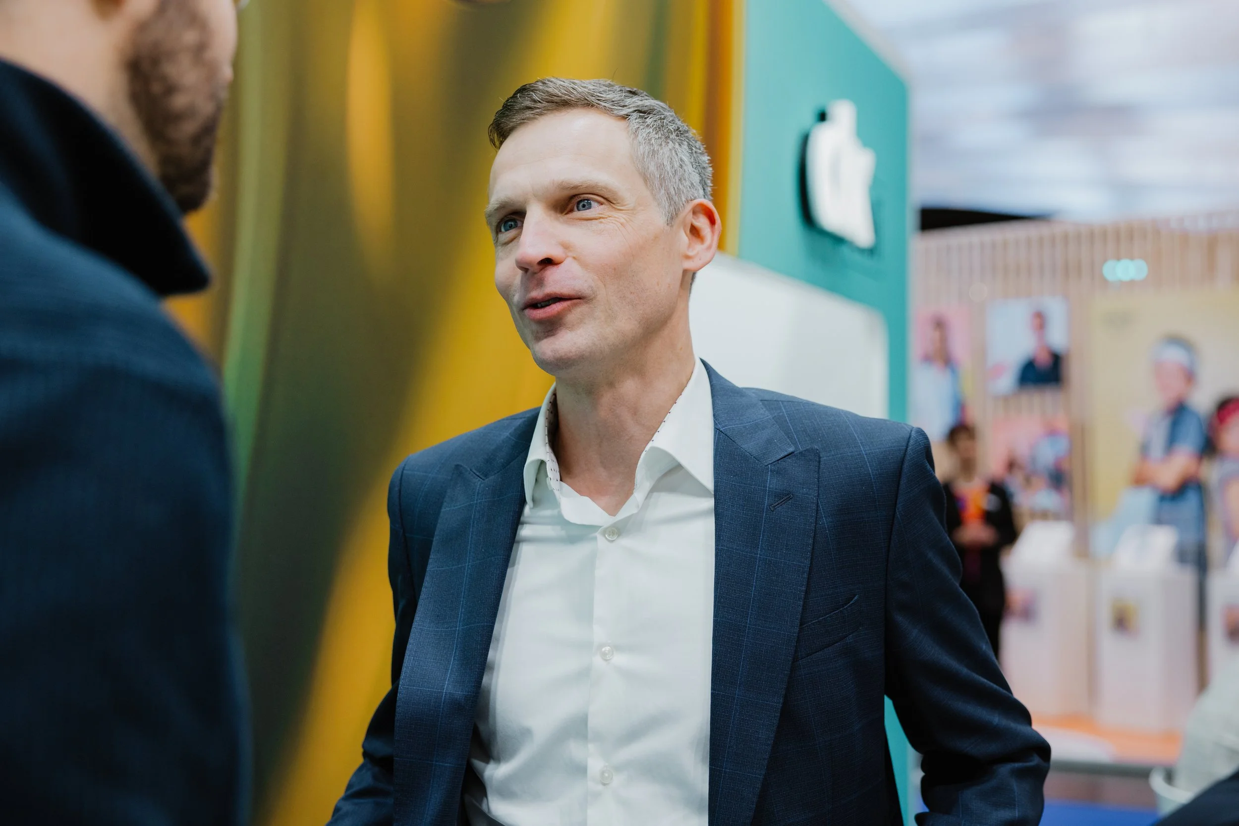 Ein Mann in einem dunklen Anzug spricht mit einem anderen Mann, im Hintergrund sind Poster und eine Präsentation zu sehen.