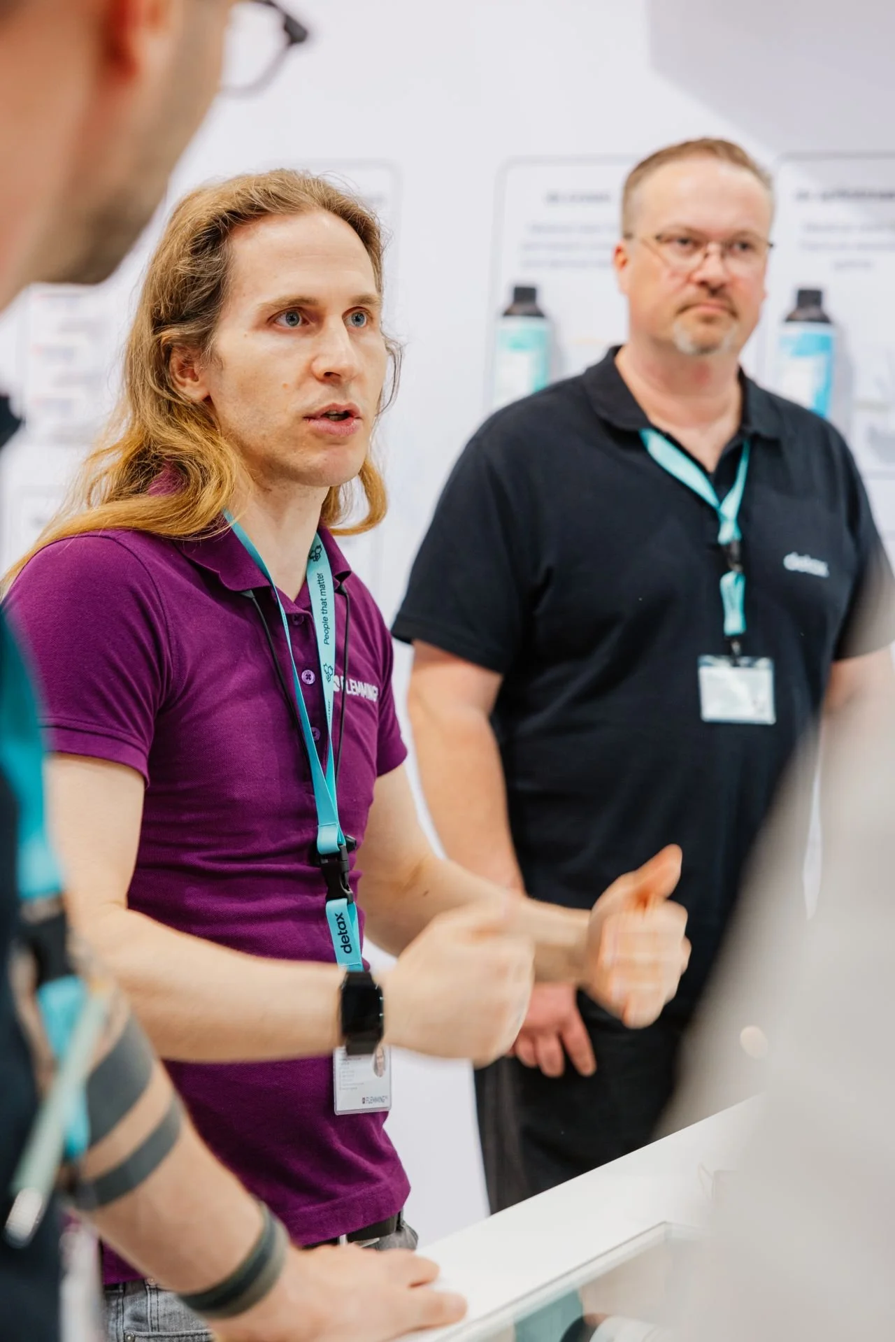 Drei Menschen bei einer Diskussion, der Mann im Vordergrund trägt ein lila Polo-Shirt, der Mann im Hintergrund trägt ein schwarzes Polo-Shirt, alle mit Namensschildern und Lanyards.